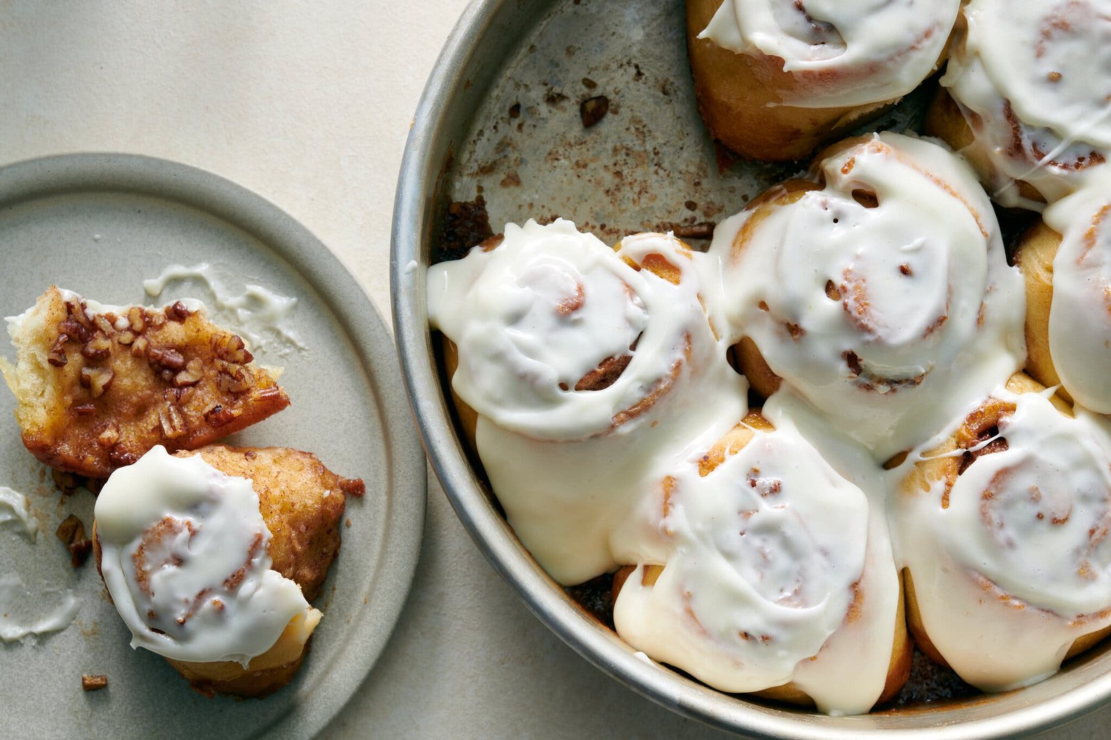 A metal pan of icing-covered cinnamon rolls has one cinnamon roll removed and served on a gray ceramic dish.