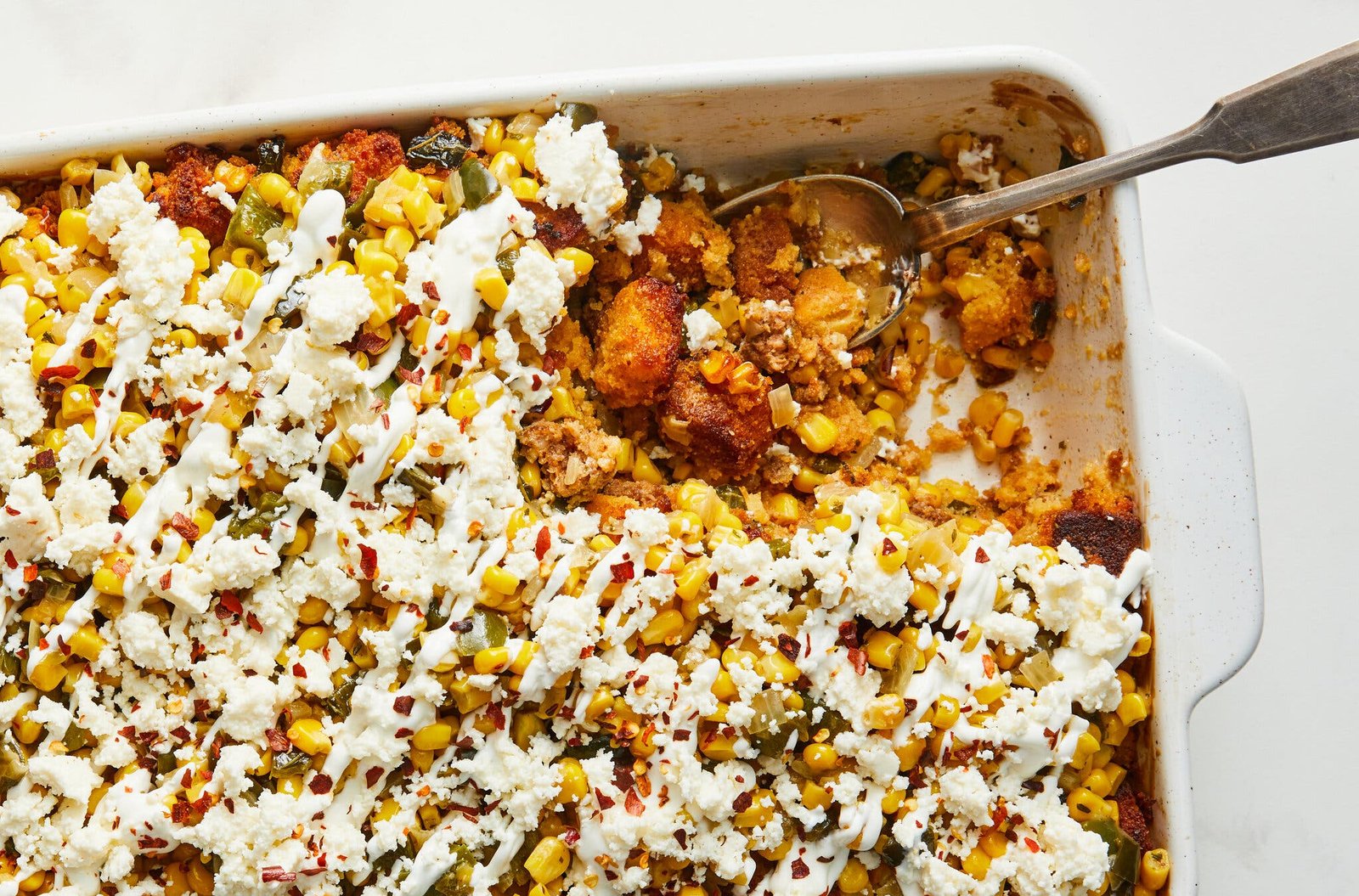 A square baking dish is filled with cornbread stuffing and topped with corn, queso and crema.