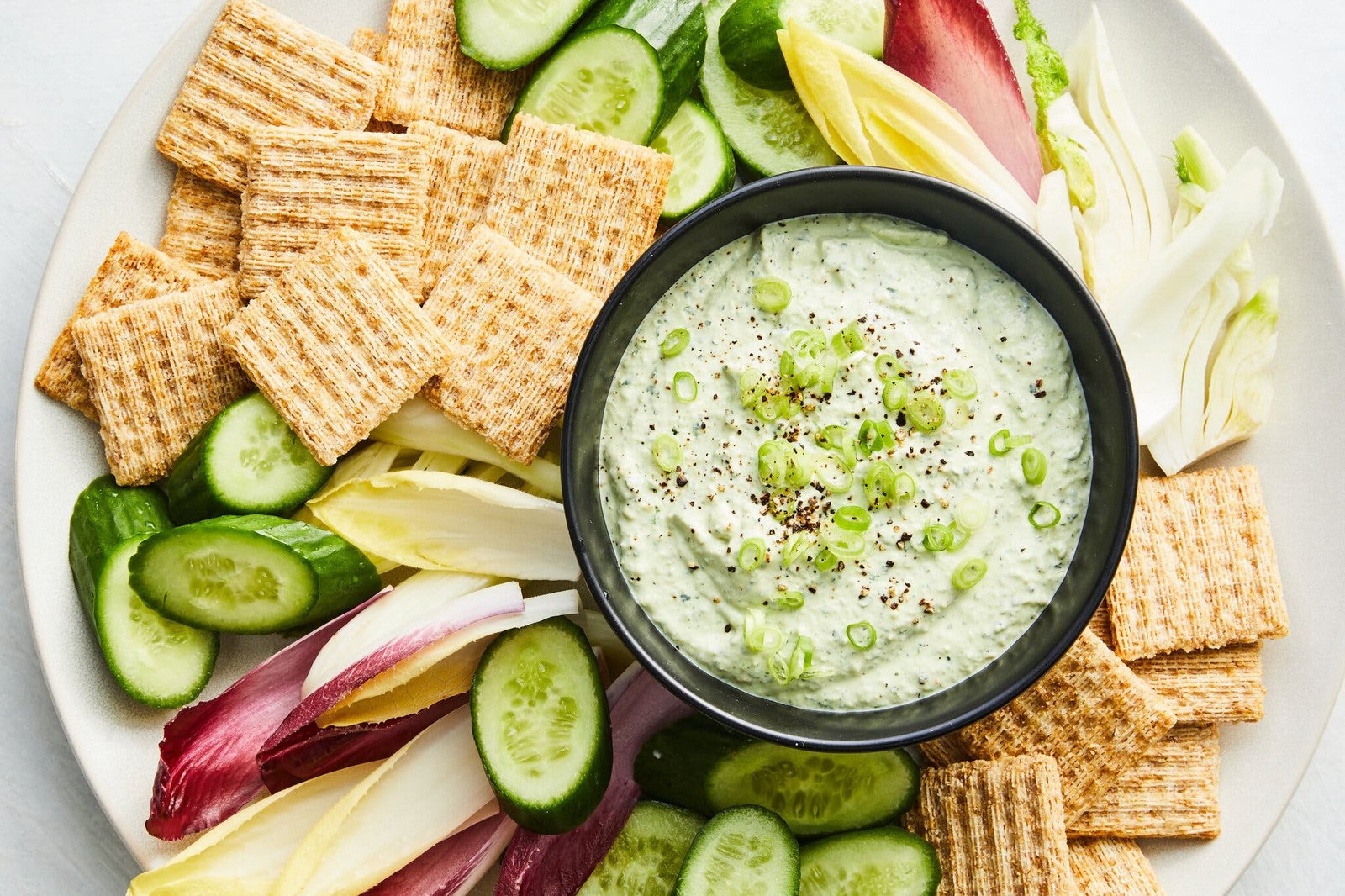 A green dip topped with scallions is surrounded by sliced vegetables and wheat crackers.