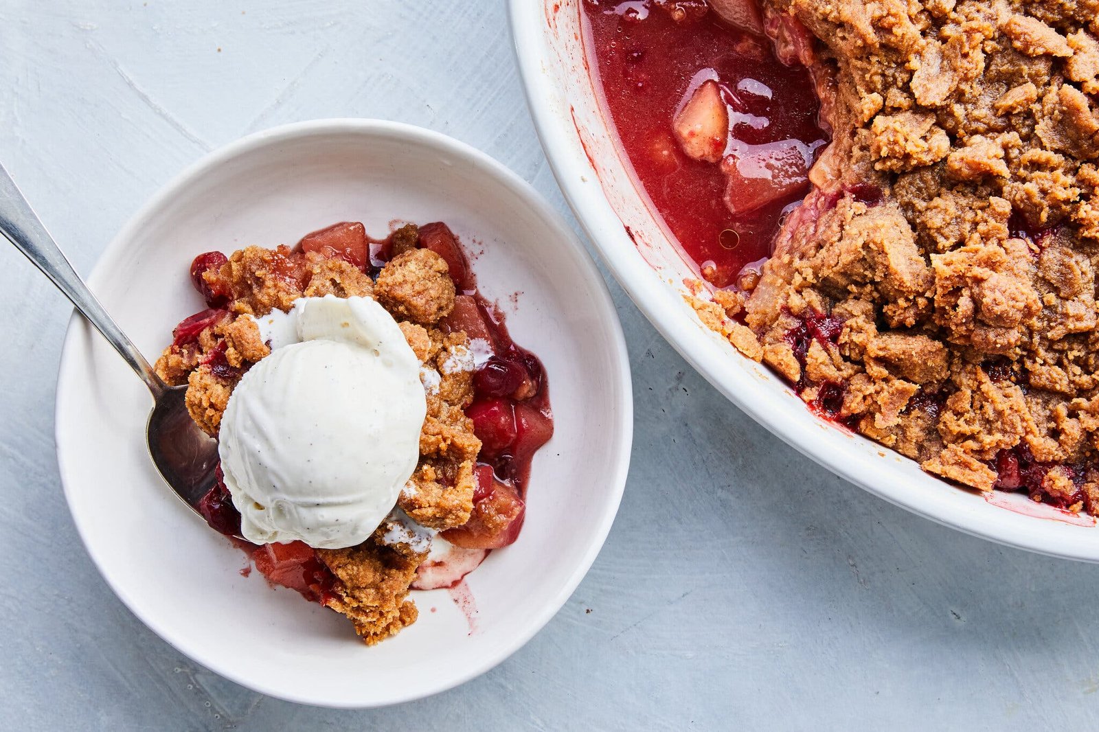 A white baking dish and bowl are filled with pears and cranberries and topped with crumb topping and ice cream.