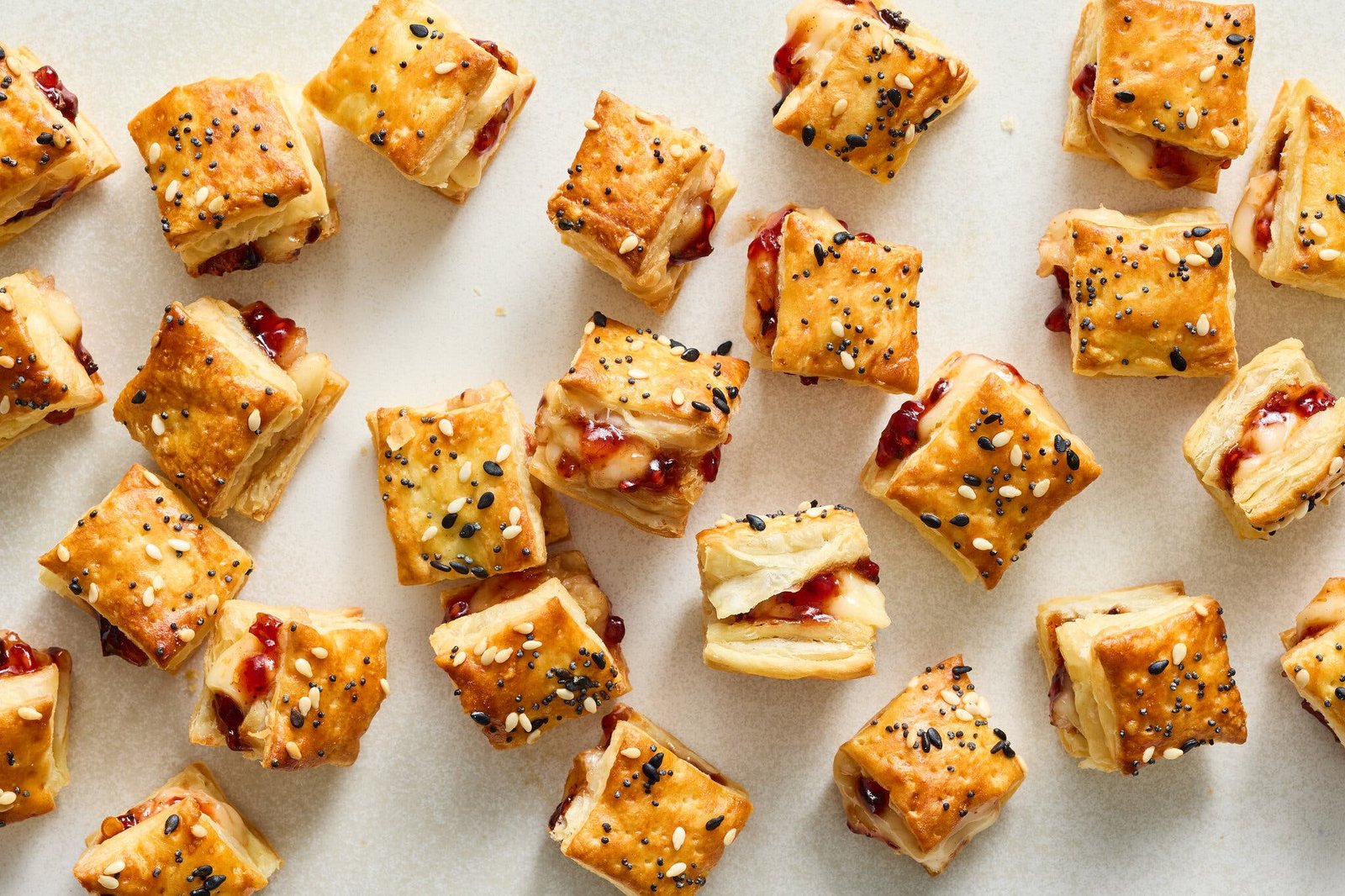 An overhead image of two dozen pastry puff squares, finished with seeds and filled with raspberry preserves and chile crisp.