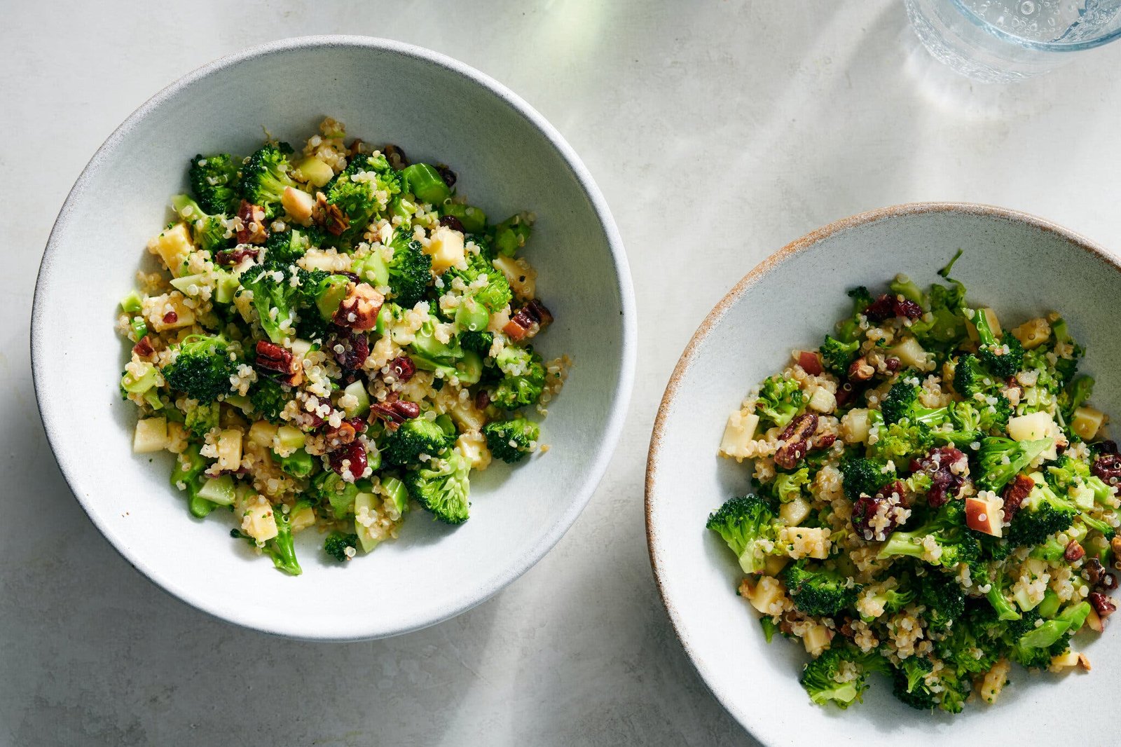 Two bowls filled with broccoli and quinoa are topped with crunchy nuts and dried fruit.