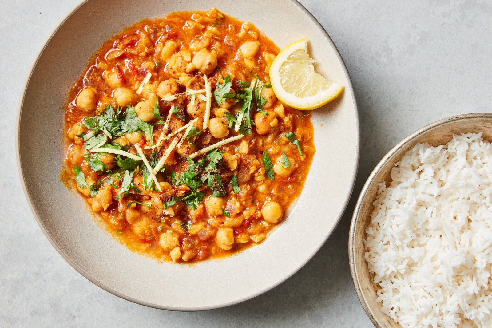 A bowl of spiced chickpeas in tomato sauce, topped with thinly sliced ginger and cilantro. A lemon wedge rests on the side of bowl. To the right sits a silver bowl with white rice.