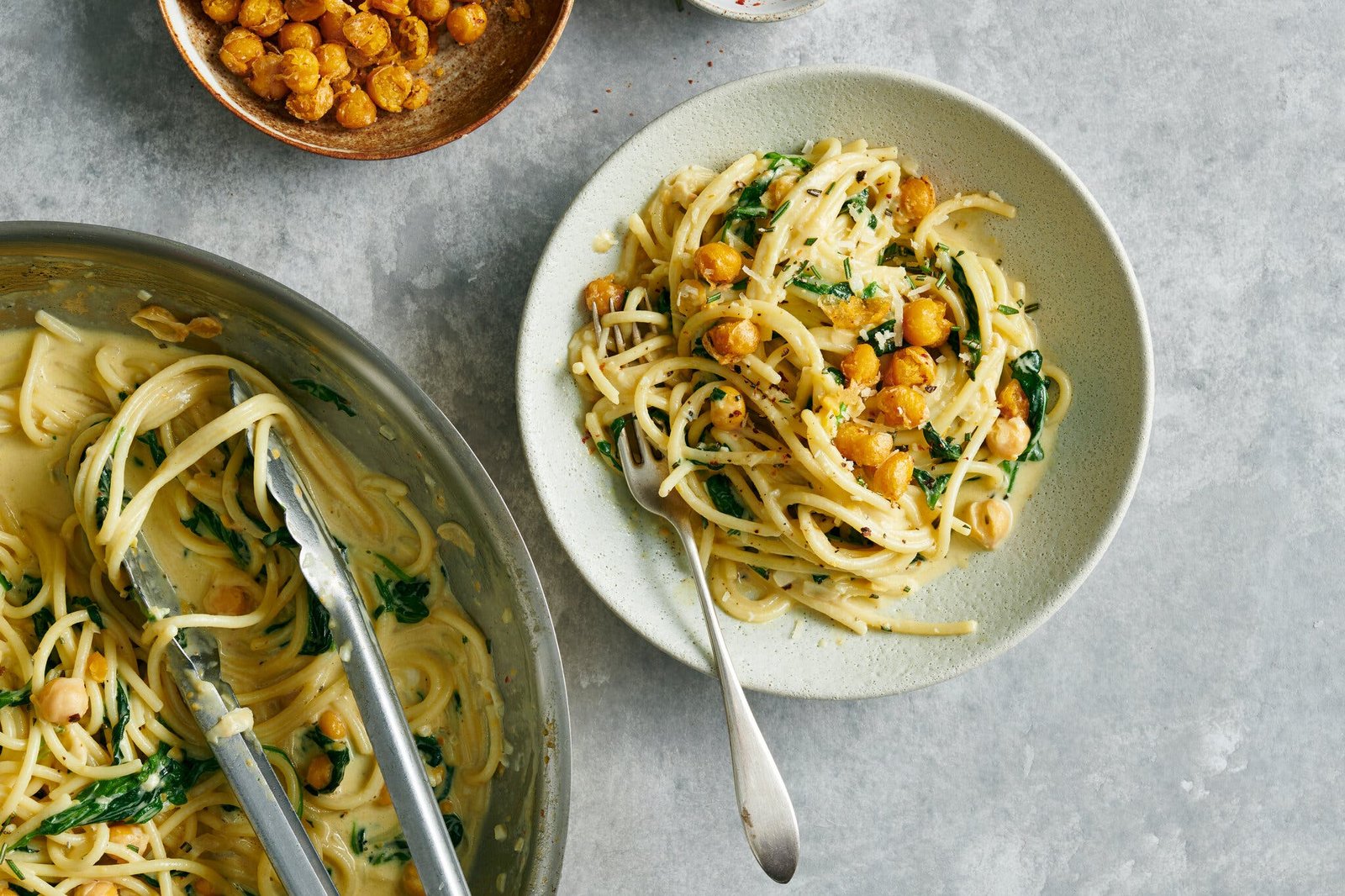 A plate of spaghetti, in a creamy sauce with chickpeas and spinach. To the left, a pan with the same pasta and a pair of tongs. 