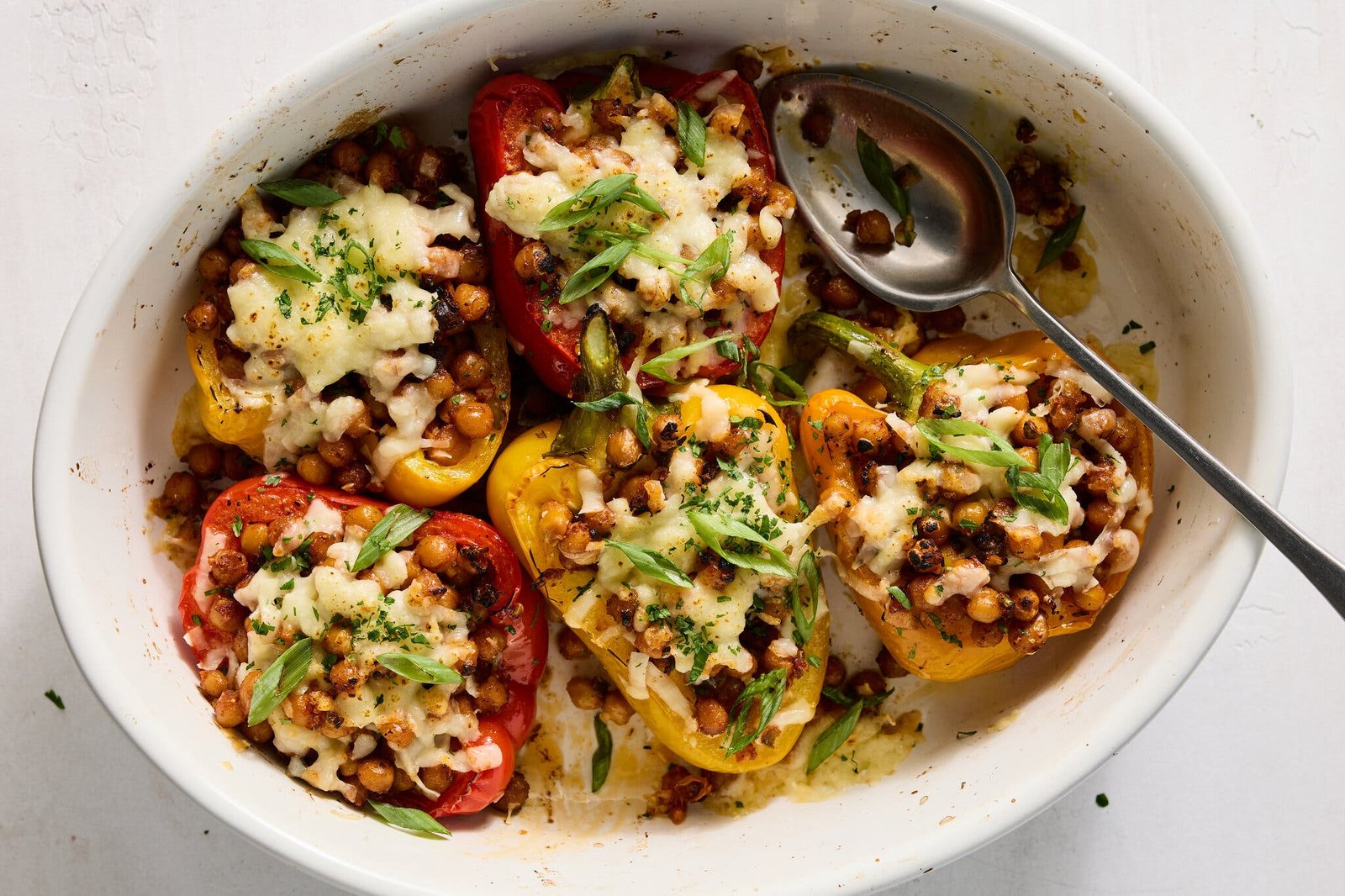 A white baking dish with five stuffed bell peppers filled with chickpeas and melted cheese, along with a silver serving spoon.