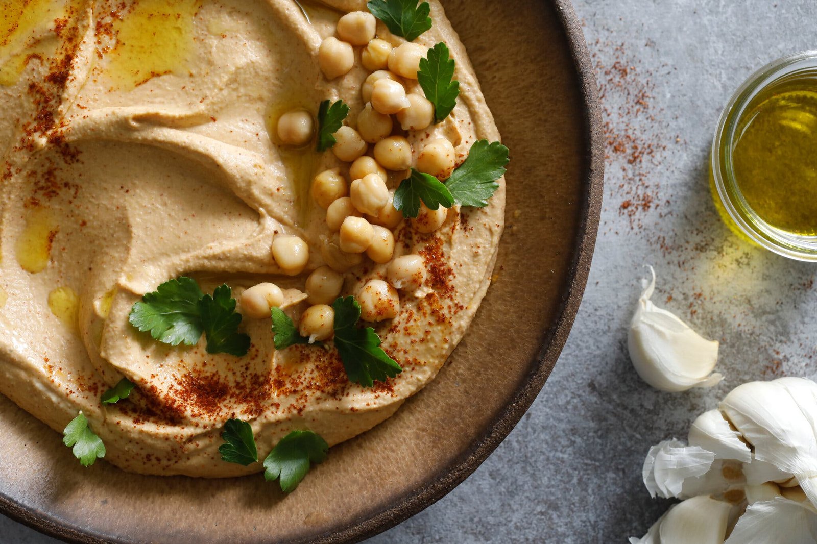 A bowl of hummus, topped with olive oil, paprika, parsley and cooked chickpeas. Beside it, a small glass bowl of olive oil and a head of garlic.