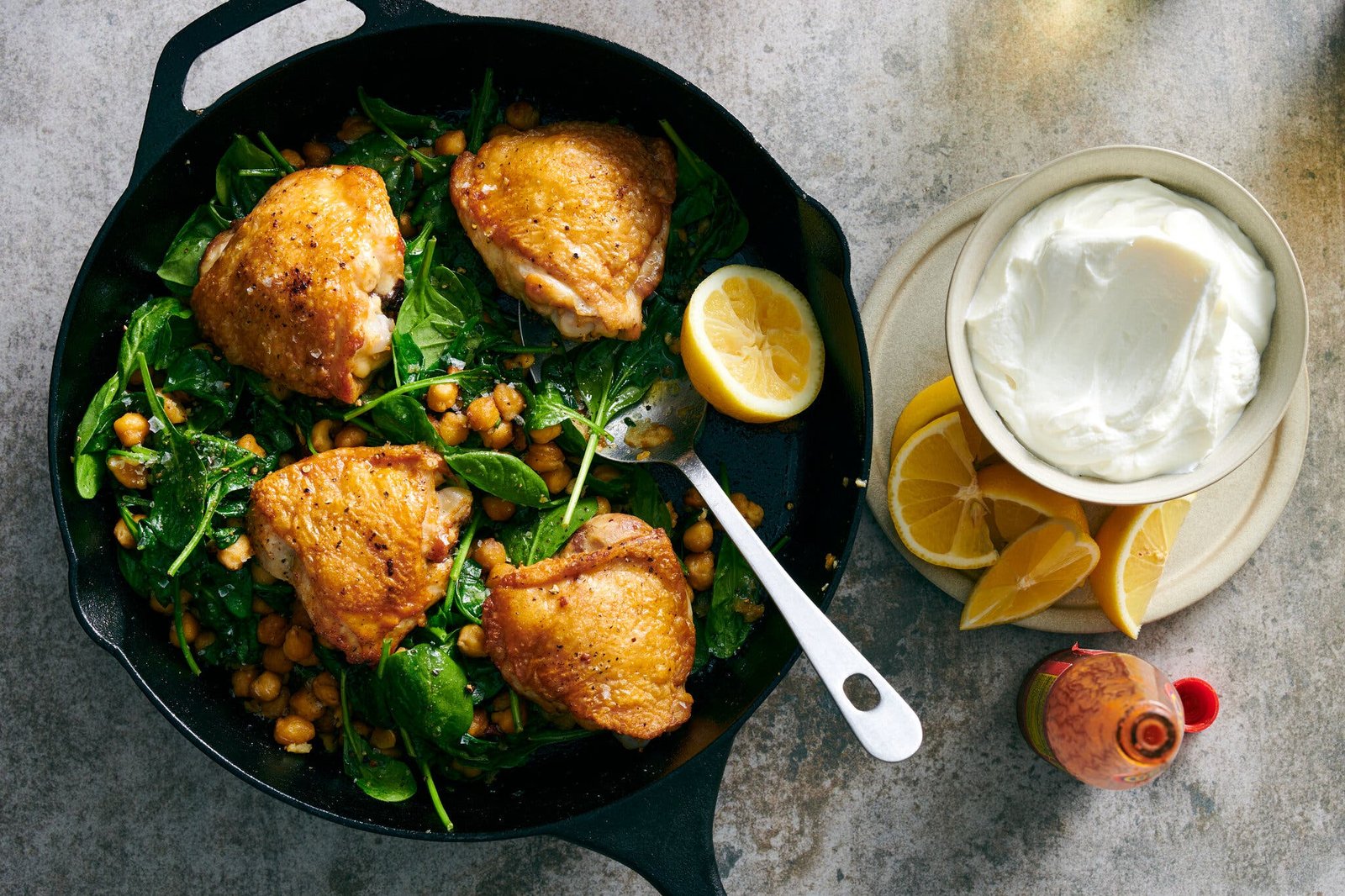 A cast-iron skillet holds crispy chicken thighs, chickpeas and baby spinach. Lemon wedges and a bowl of yogurt sit on the side.