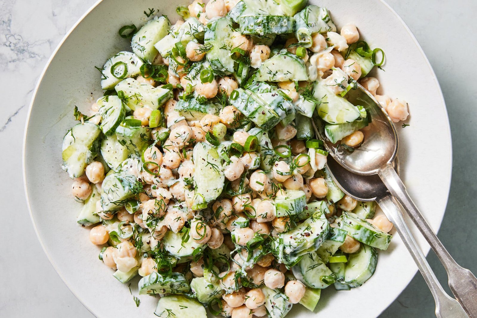 A bowl of salad with cucumbers, chickpeas, dill, mint and scallions tossed in a Greek yogurt dressing, along with two serving spoons.