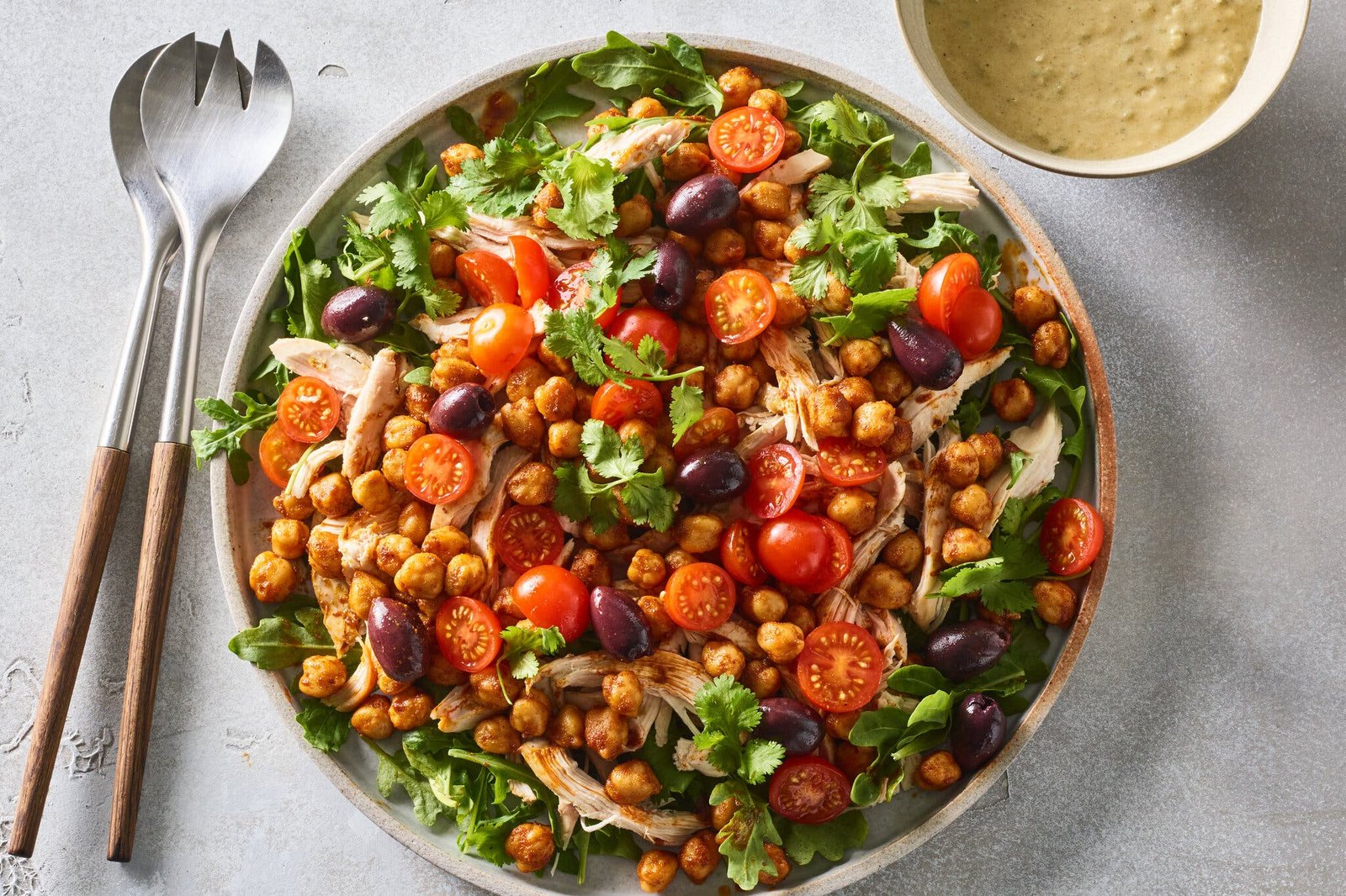 A large bowl of salad with arugula, halved cherry tomatoes, Kalamata olives, shredded chicken and cilantro. Beside it, a pair of serving utensils and a bowl of green harissa dressing.