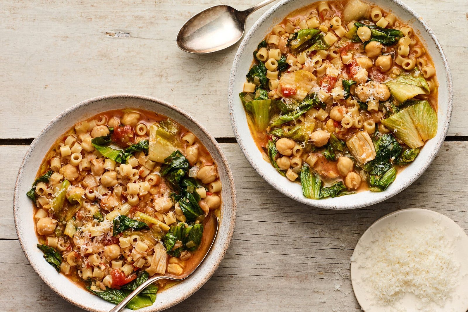 Two bowls of stew, on a wooden surface, with chickpeas, escarole, tomatoes and grated pecorino. 