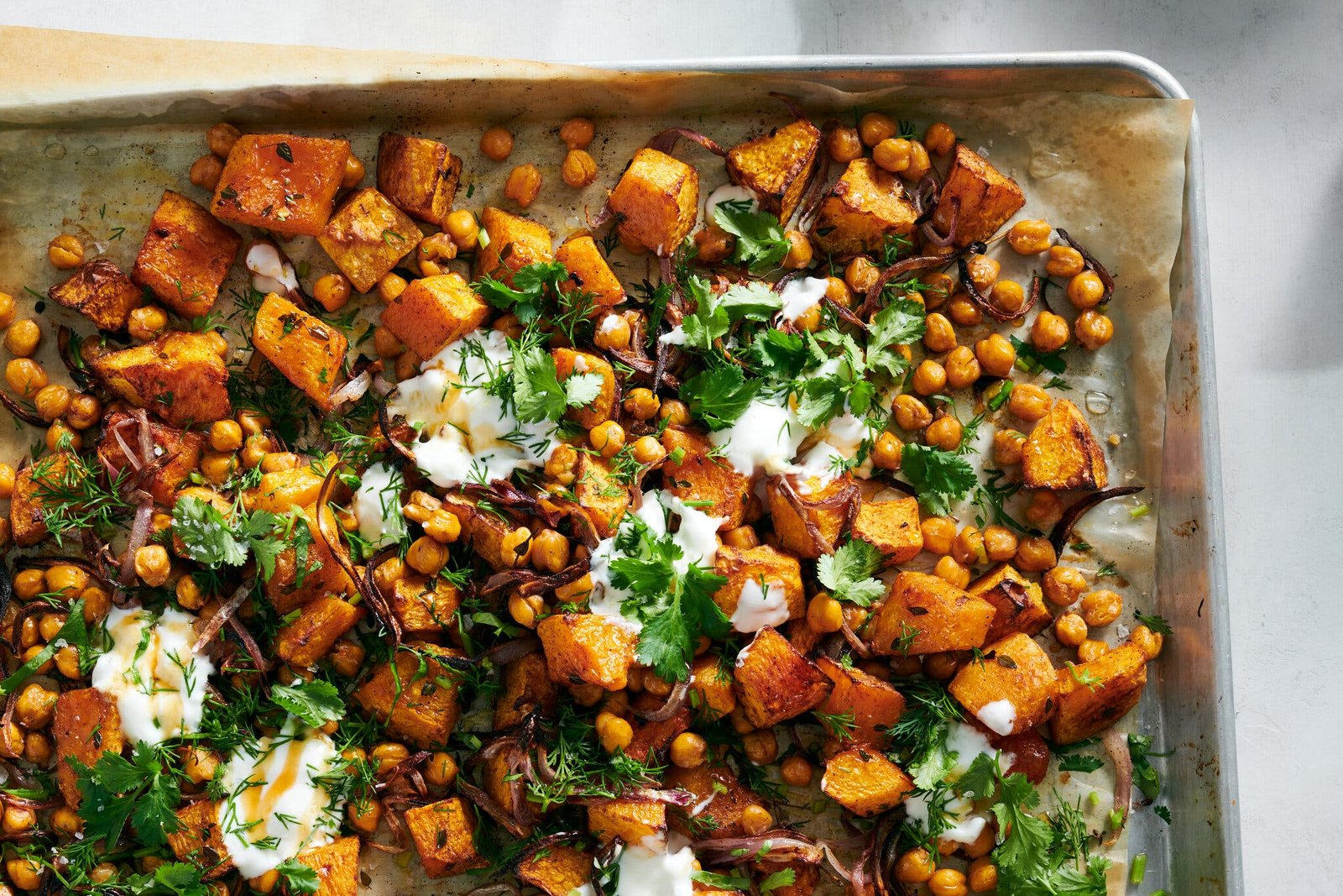 A sheet pan full of roasted squash and chickpeas sprinkled with herbs and yogurt is shot from above. A sheet of parchment paper peeks out from the bottom of the pan.