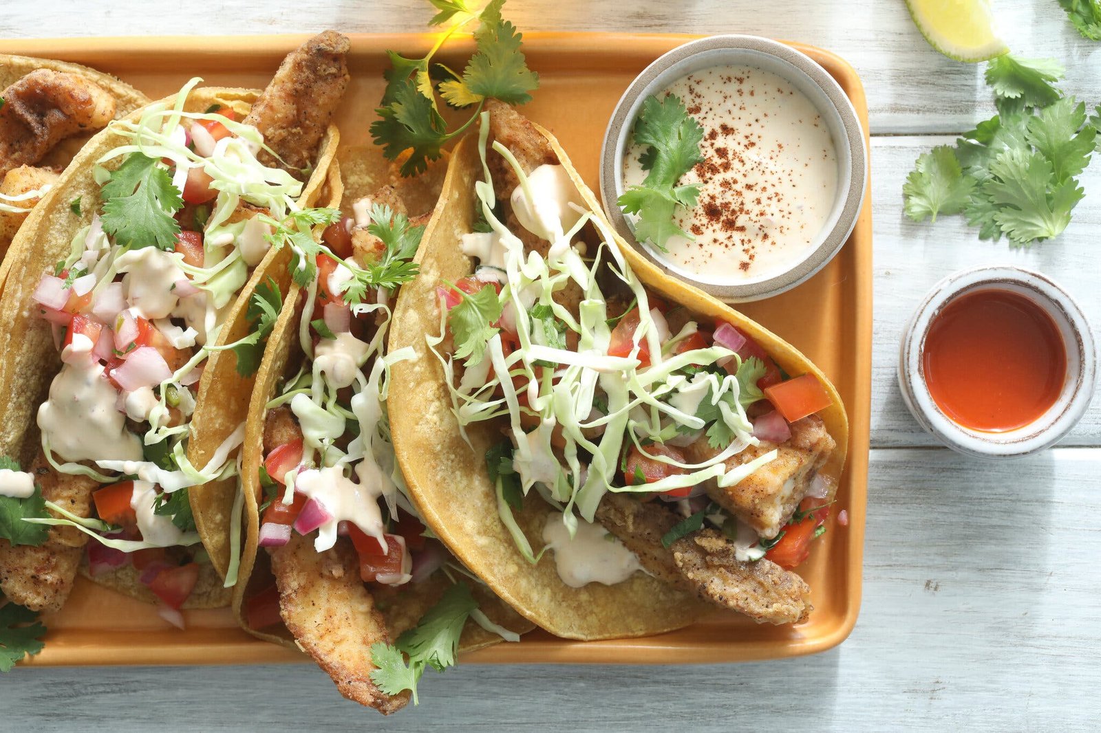 An overhead image of fish tacos on a platter topped with cabbage. Crema and hot sauce sit off to the side.