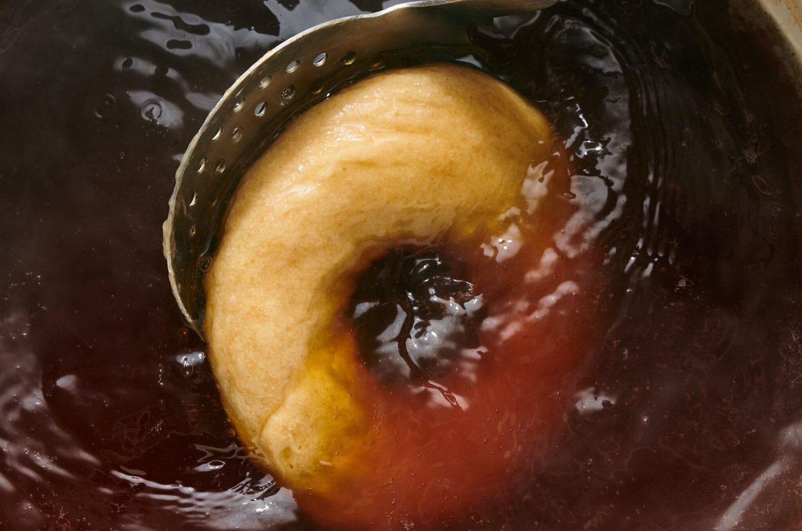 An overhead image of bagel dough being lowered into a pot of boiling water.