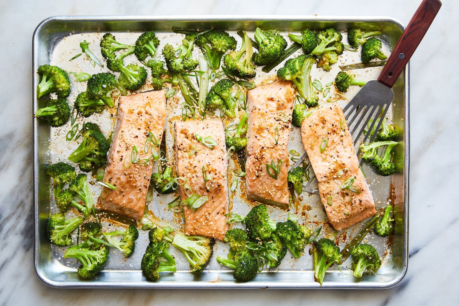 Four roasted fillets of salmon and broccoli florets are on a silver sheet pan with a fish spatula for serving.