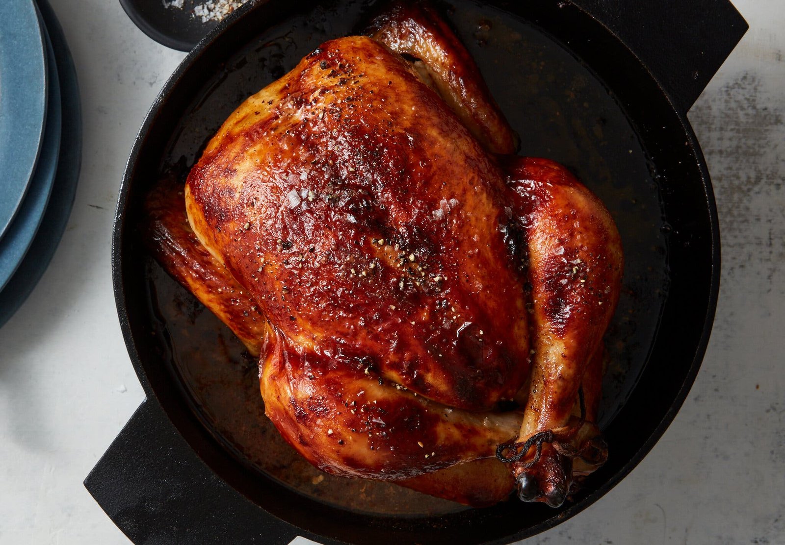 A deeply burnished buttermilk-brined roast chicken is shown in a cast-iron pot with a stack of blue plates and a small dish of salt and pepper nearby.