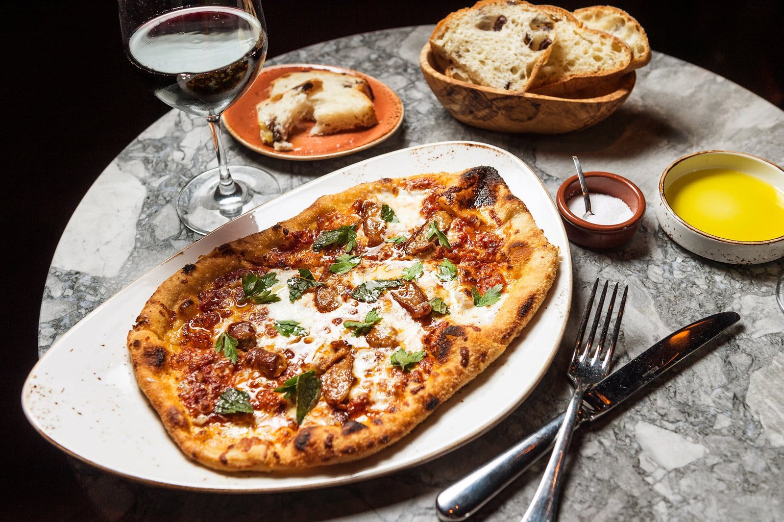 A flatbread pizza on a white plate, surrounded by a glass of red wine, bread and small bowls of salt and oil.