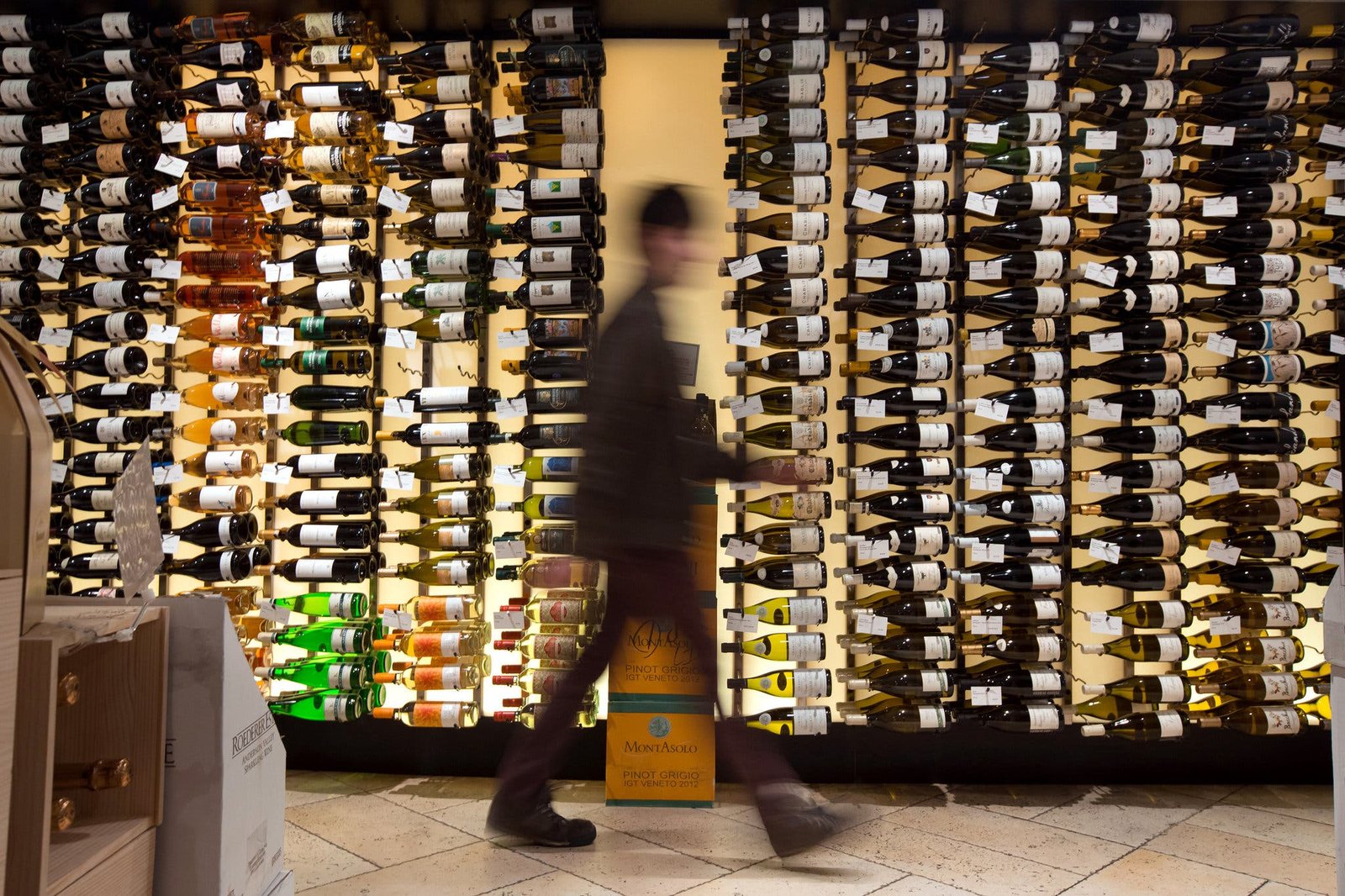 A blurred figure walks in front of a brightly lit wall densely packed with wine bottles.