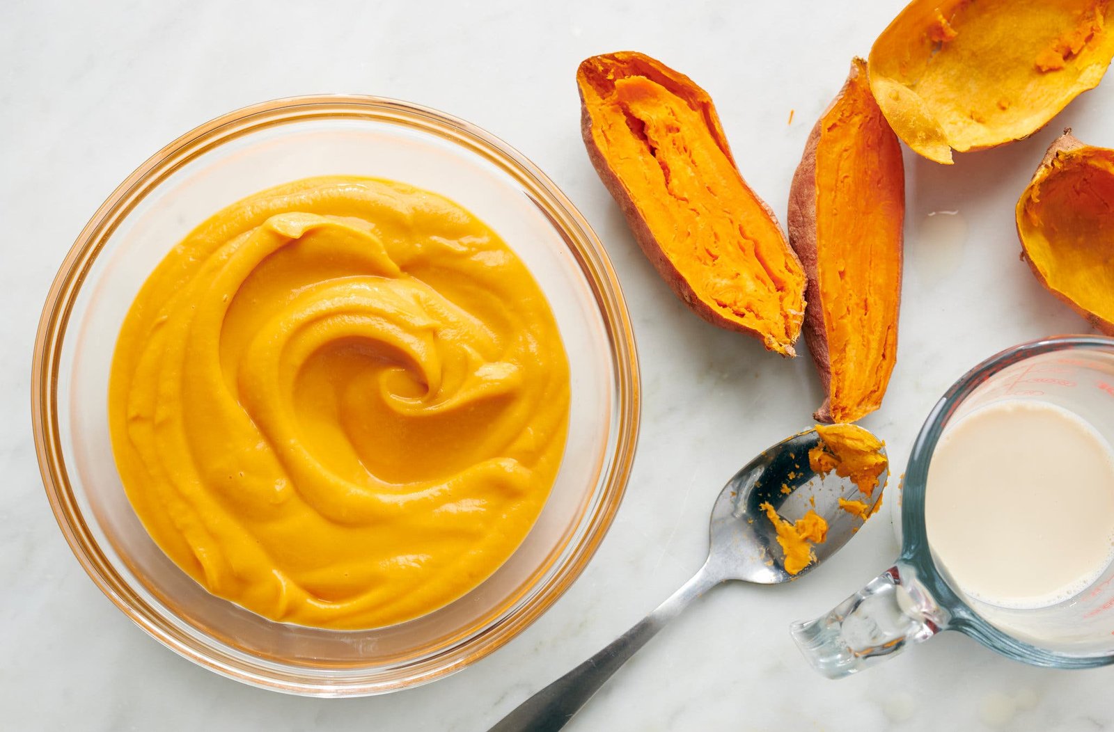 Yellow purée in a glass bowl, with sweet potato halves and a measuring cup with milk nearby.