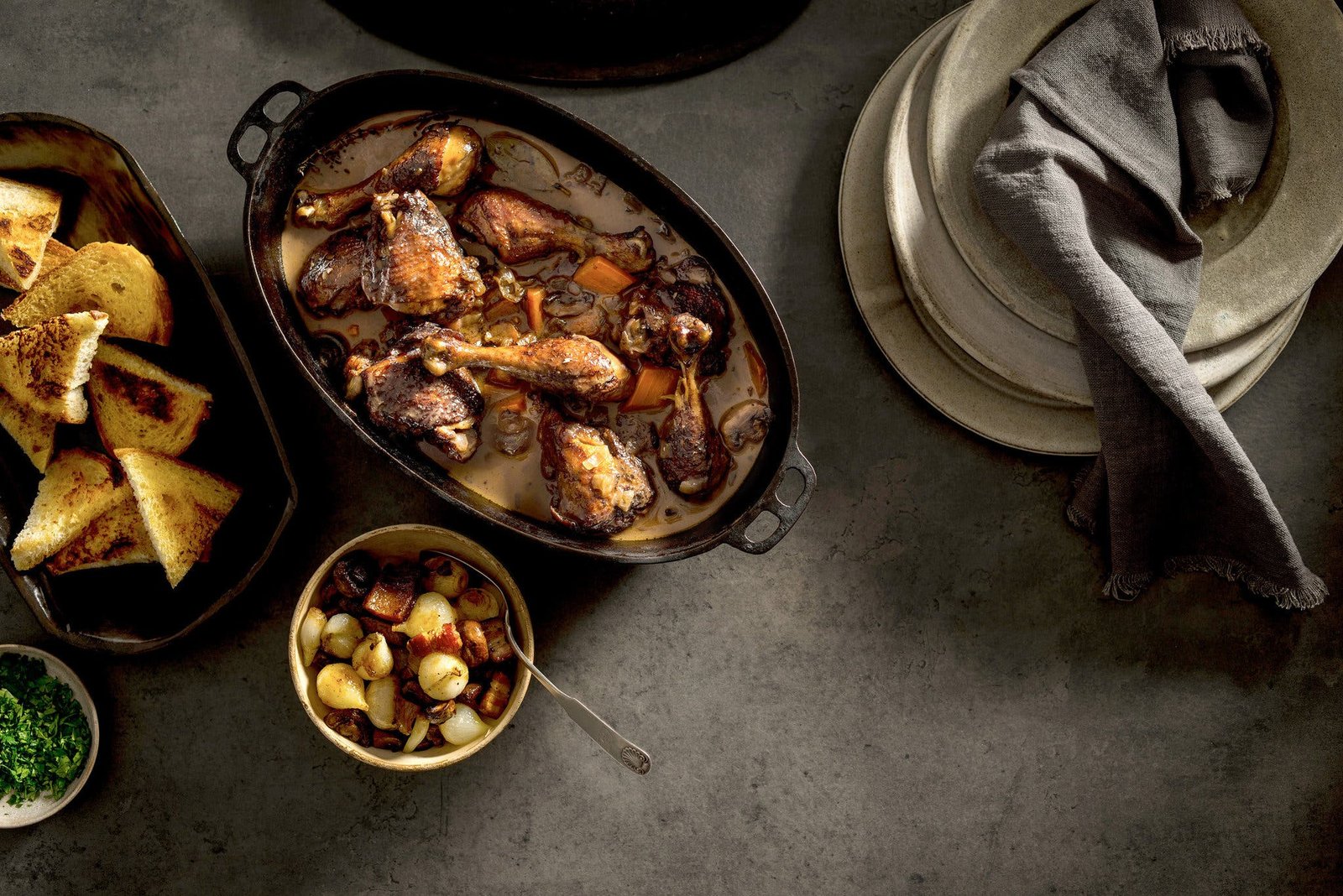 An overhead image of a dutch oven filled with coq au vin, next to a platter with bread and a pile of plates.