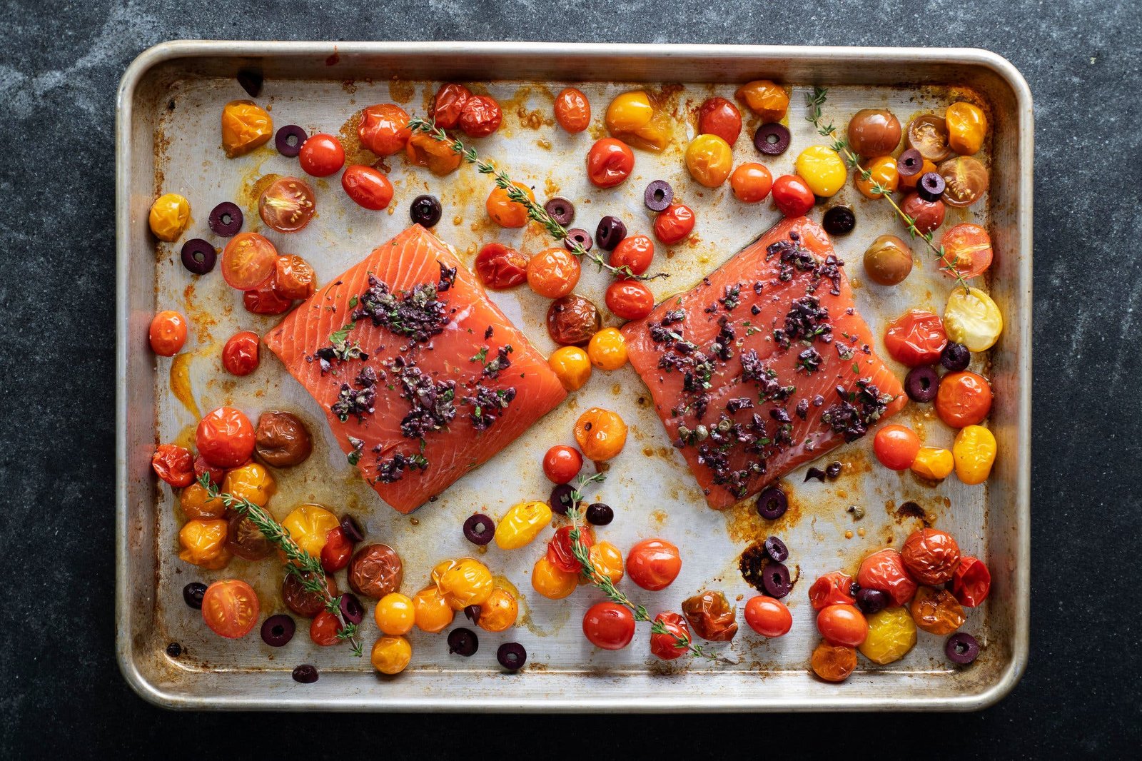 An overhead image of raw salmon on a sheet pan surrounded by blistered tomatoes and olives.