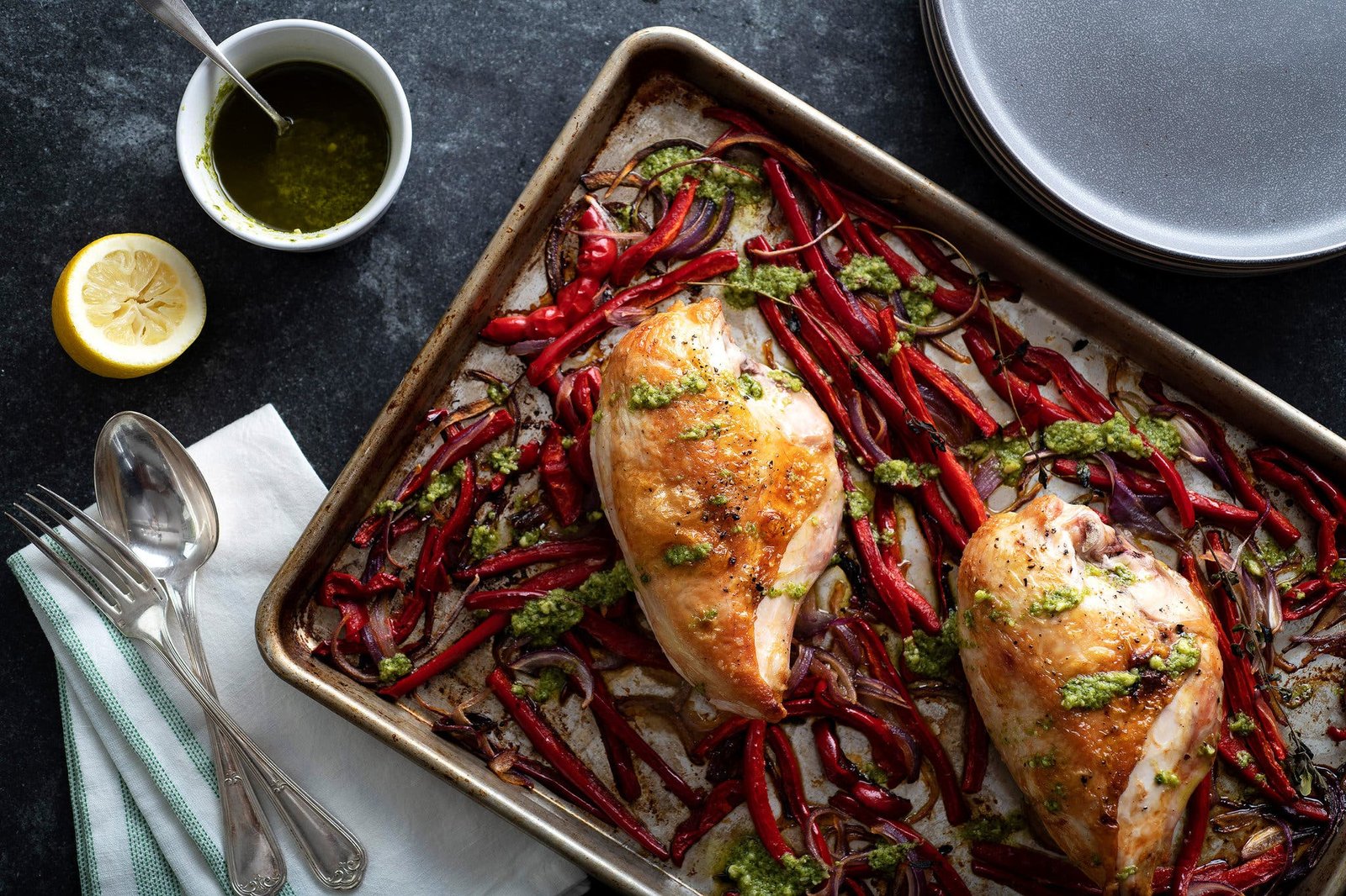 An overhead image of chicken breasts and red peppers on a sheet pan next to a pesto and lemon.