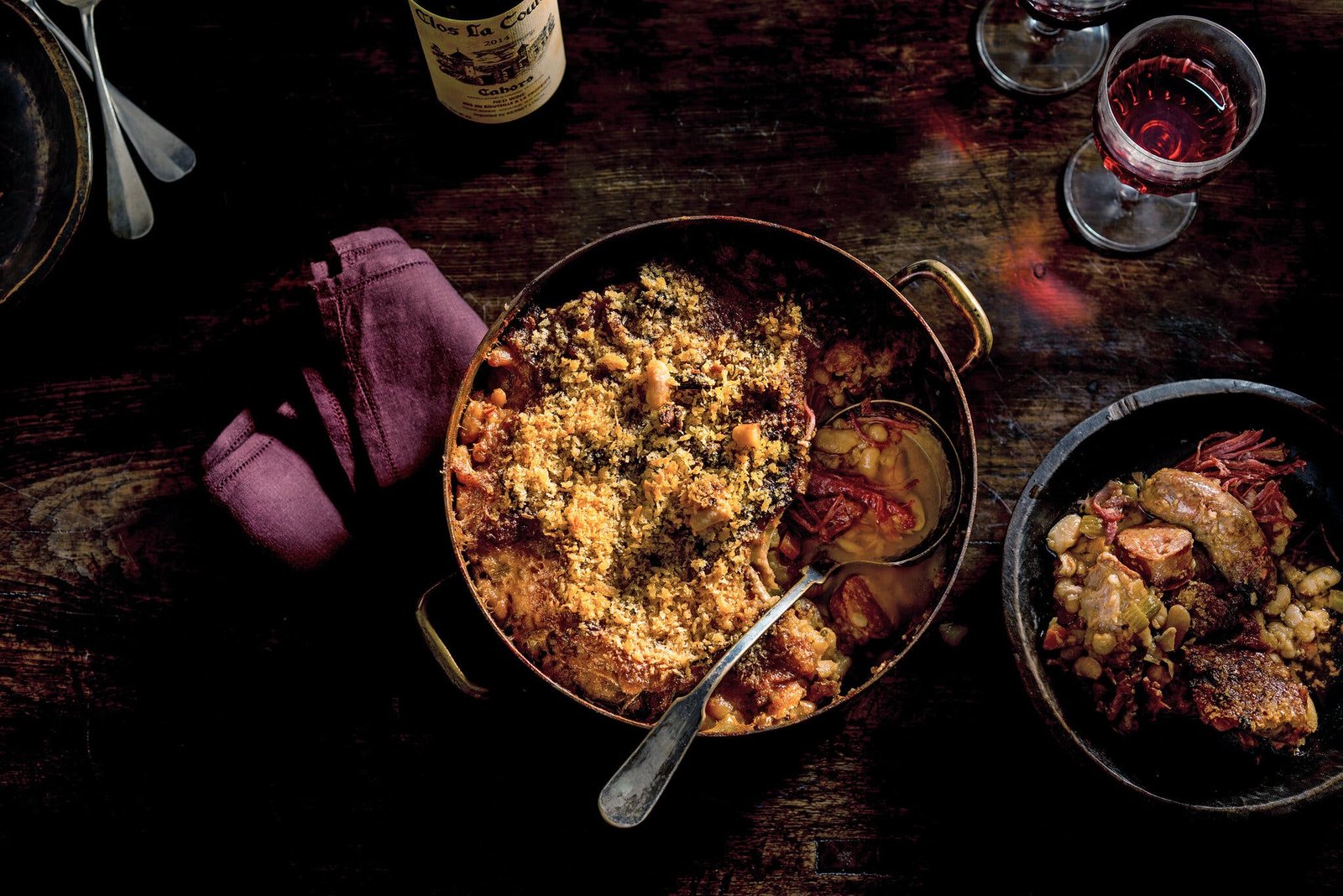 An overhead image of cassoulet in a pot next to a single serving of the dish.