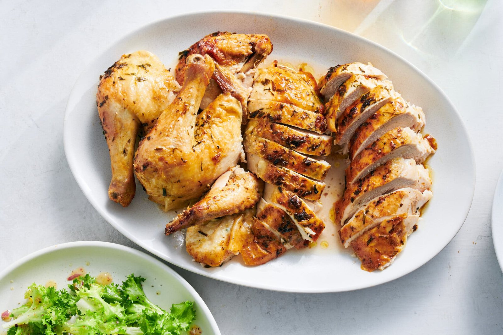 A white oval platter holds pieces of a carved chicken. Below it, a plate with salad peeks in.