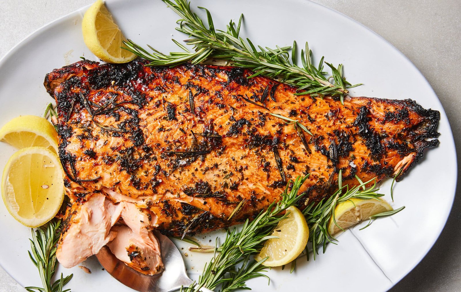 A large salmon fillet with lemon-herb marinade is on a white platter with rosemary sprigs and lemon wedges.