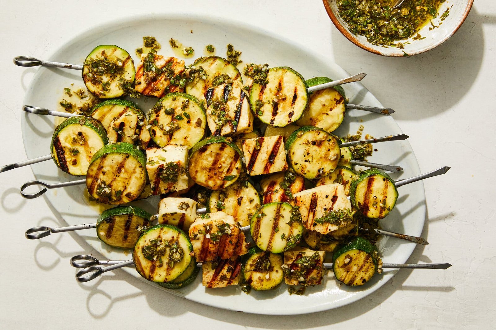 A white platter holds skewers of grilled halloumi and zucchini with salsa verde.