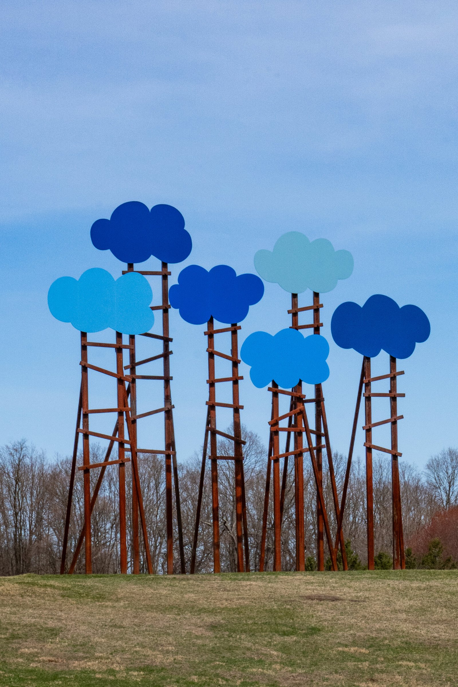 Six powder-coated aluminum cloud shapes, painted in various shades of blue, are perched more than 30 feet atop giant ladderlike steel supports. They're outside on dry grass. 