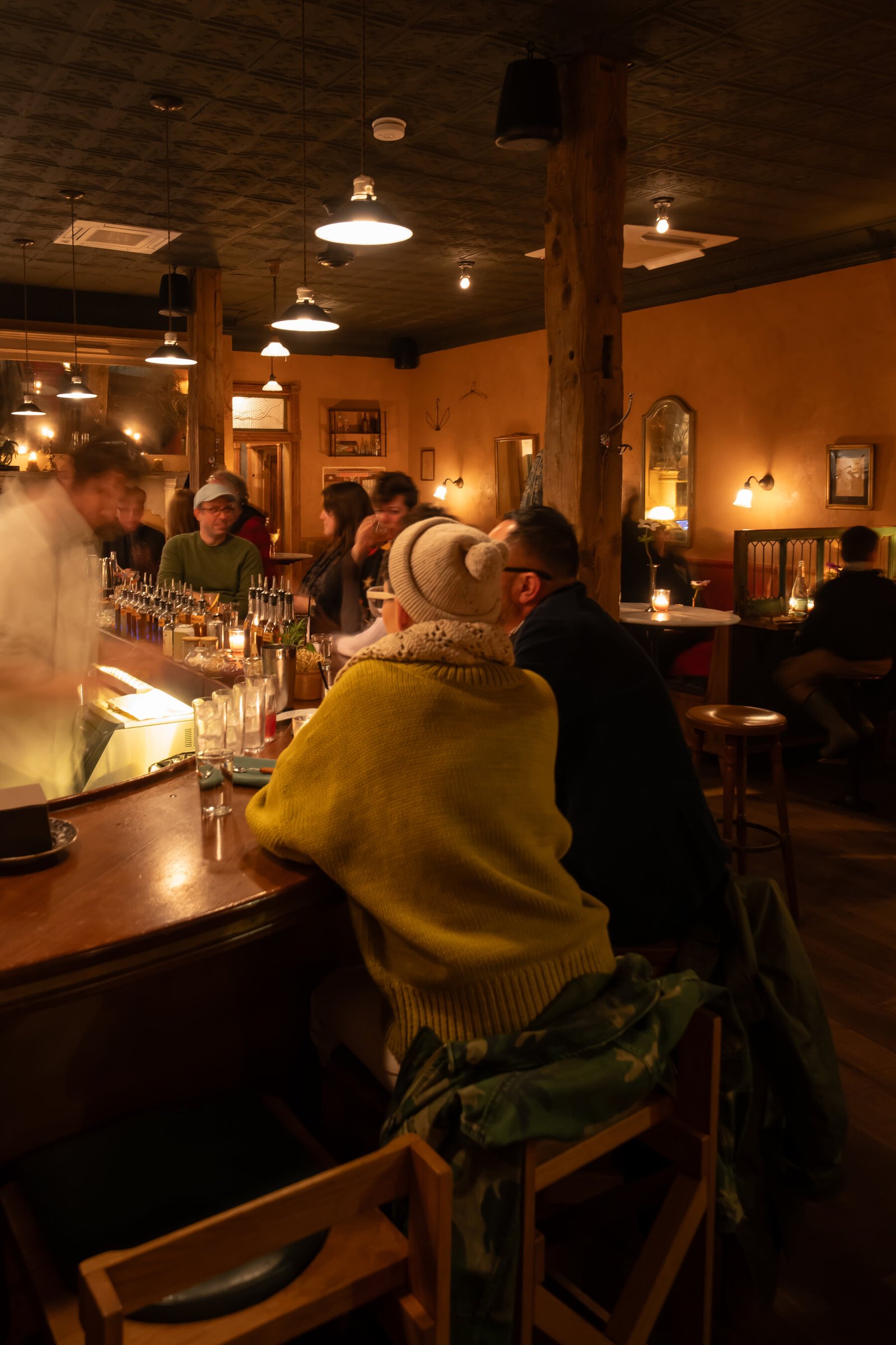 The interior of a bar, with patrons seated at the bar and a bartender behind it. The atmosphere looks warm and inviting. 