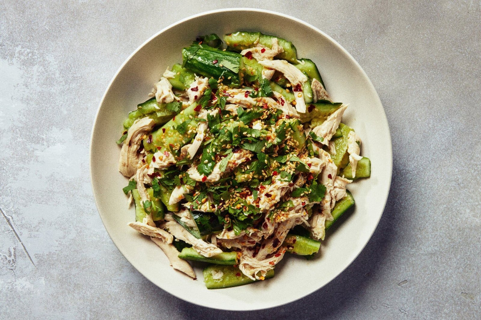 A beige bowl holds a salad of shredded chicken, sliced cucumbers and green herbs, sprinkled with crushed red pepper.