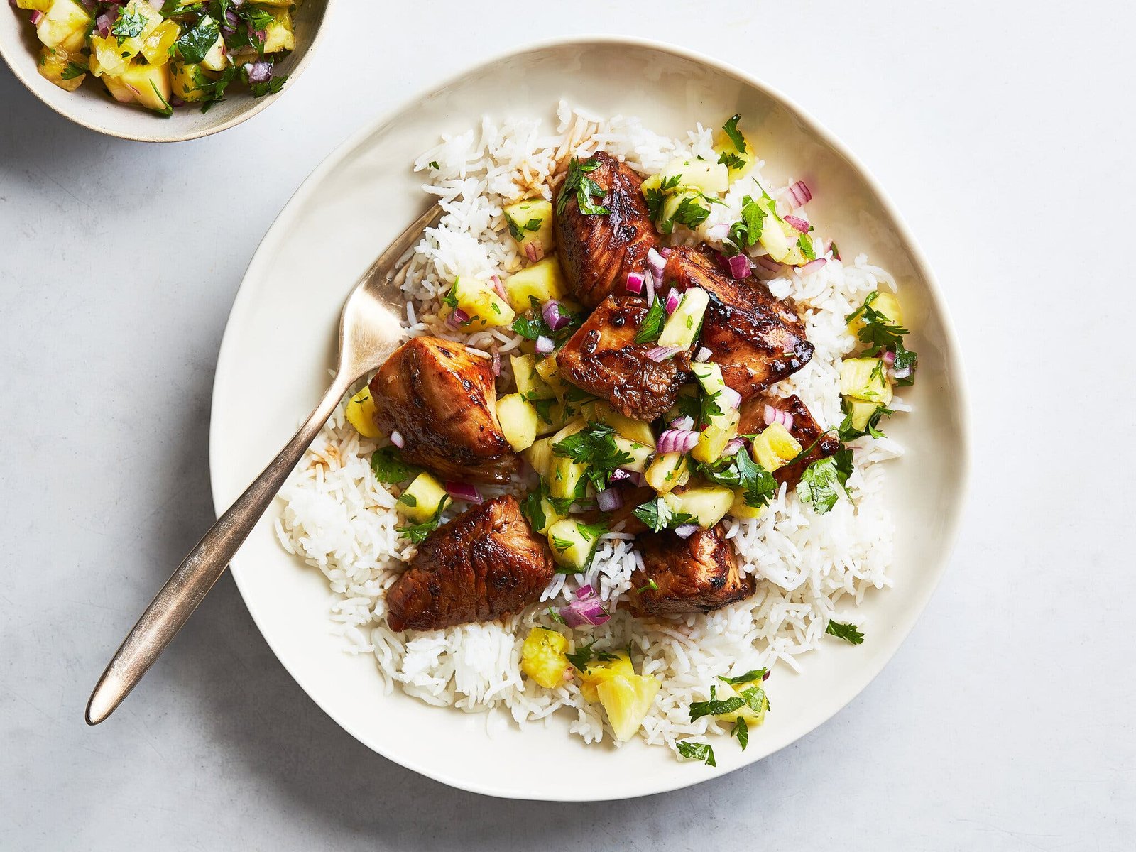 A white plate holds grilled chicken pieces over white rice, topped with pineapple, red onion and cilantro. A fork rests on the plate.