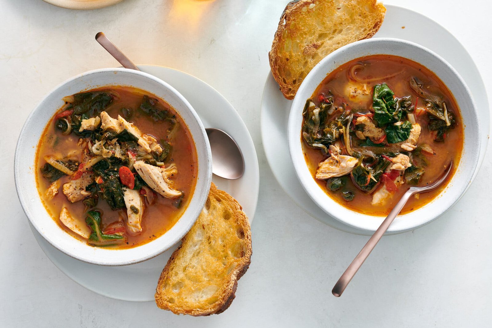 Two white bowls of a ruddy chicken soup with greens sit on white plates, each with a piece of toasted bread leaning against the bowl and a spoon resting inside.
