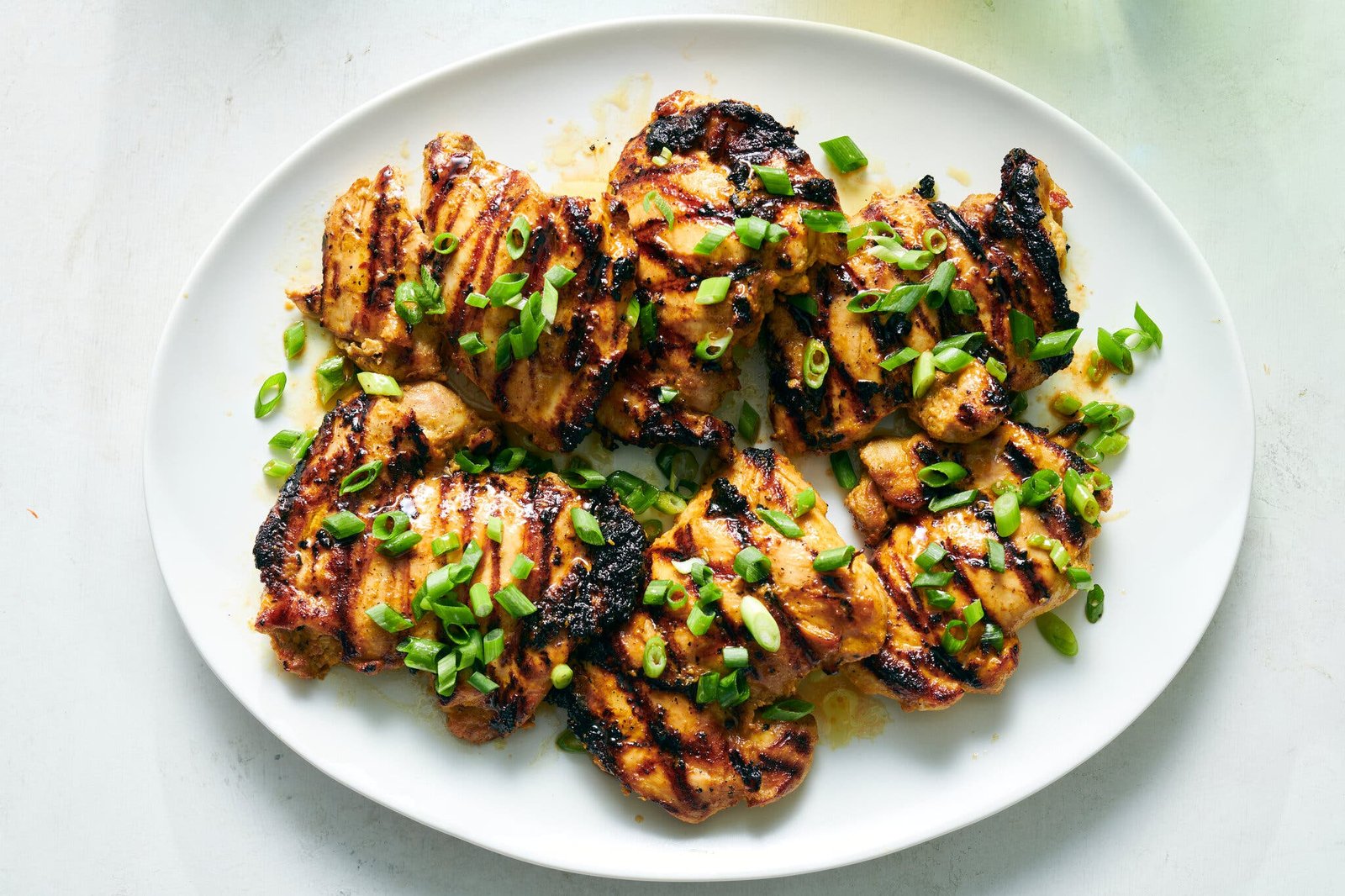 Grilled chicken thighs with visible grill marks are arranged on a white plate and topped with chopped scallions.