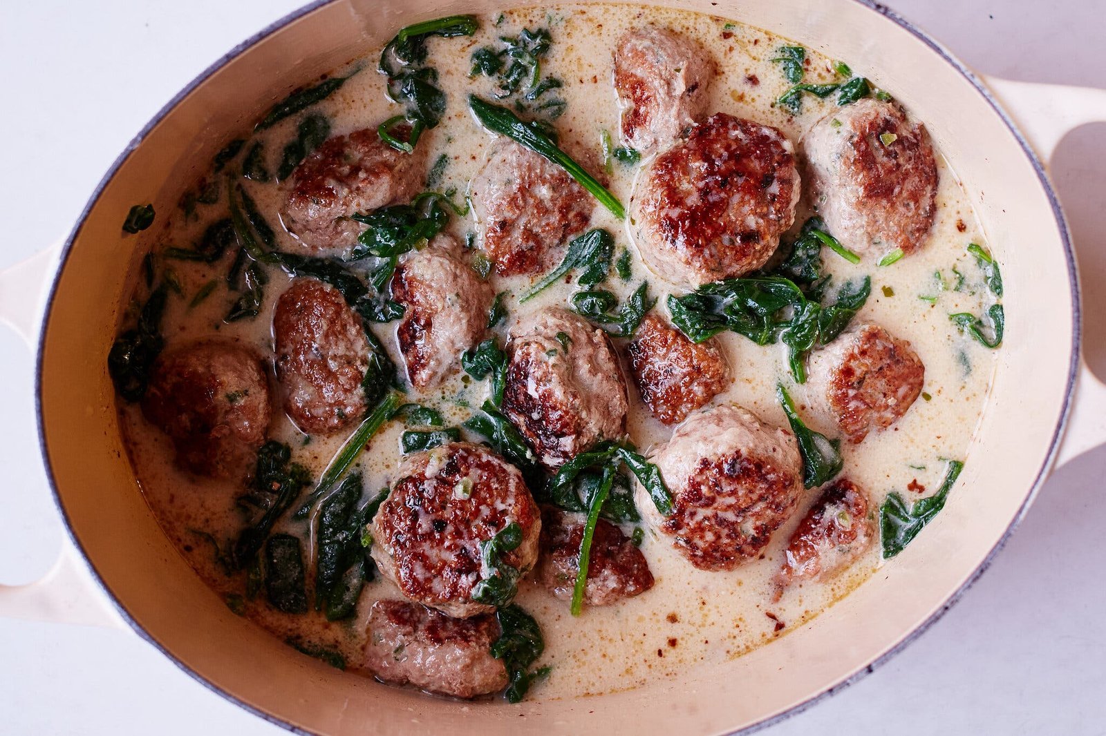 A creamy soup with browned meatballs and vibrant green spinach leaves in a light-colored pot.
