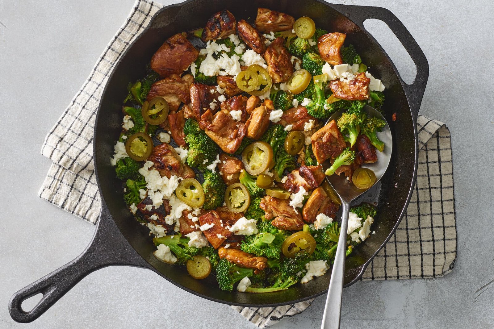 A cast iron skillet holds spicy honey chicken with broccoli, crumbled white cheese and sliced jalapeños.