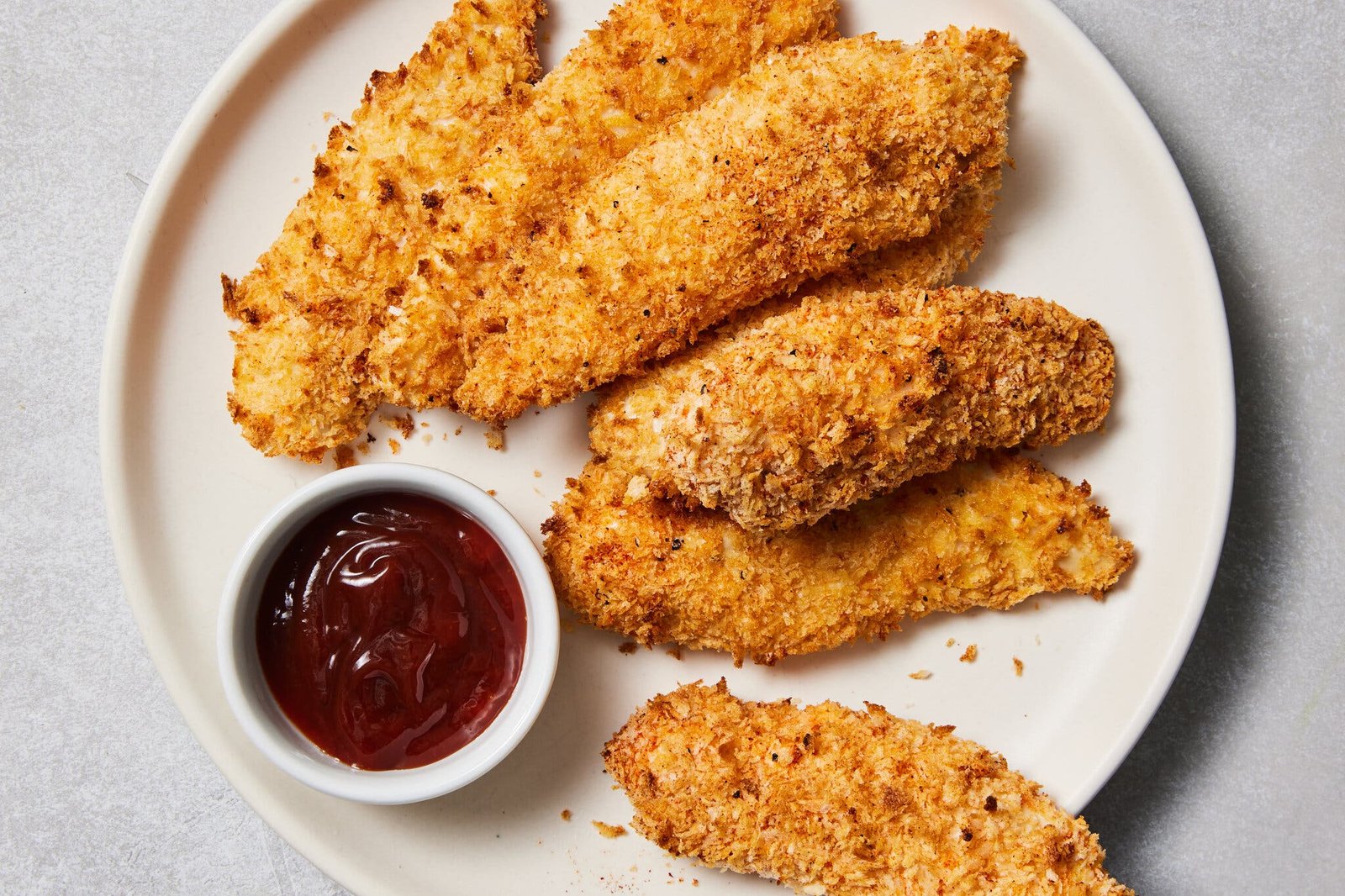 Golden-brown air fryer chicken tenders are arranged on a white plate next to a small bowl of red dipping sauce.