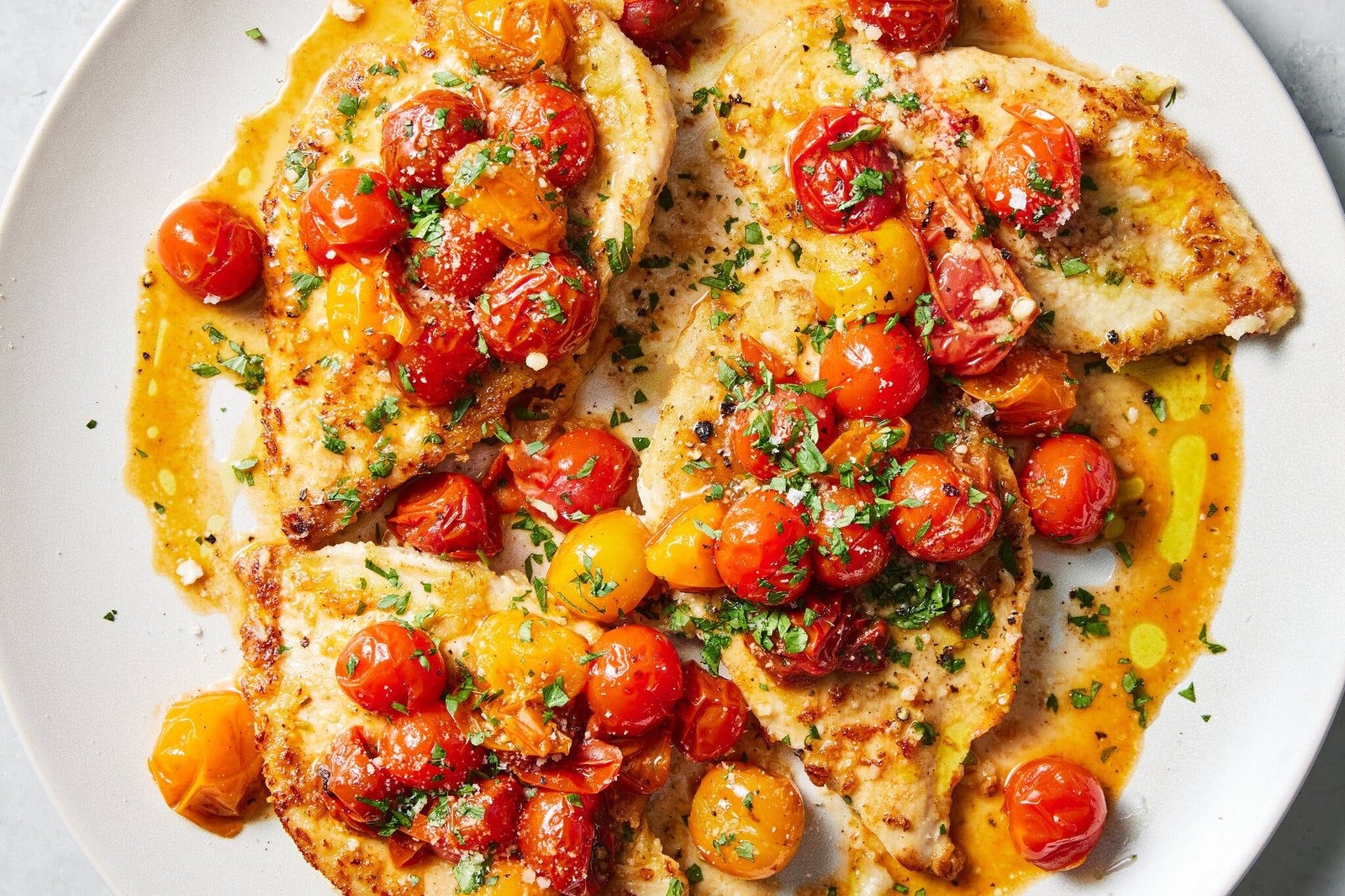 Four pieces of lemon and garlic chicken topped with red and yellow cherry tomatoes and herbs on a white plate.