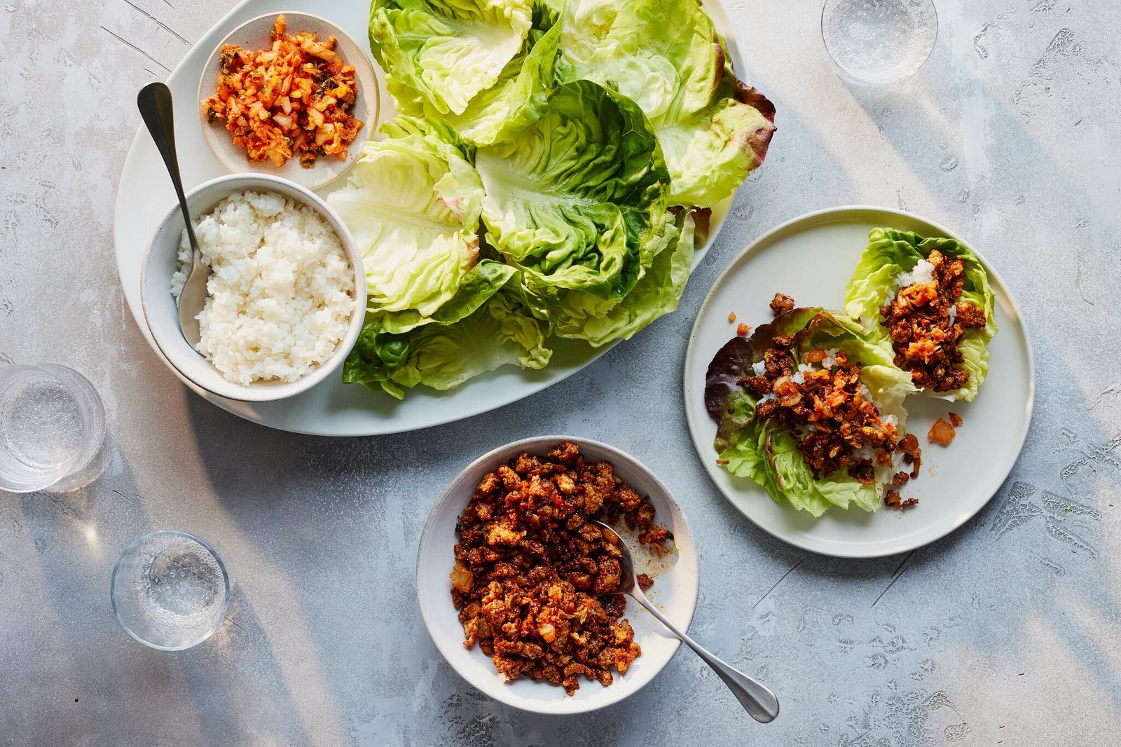 A platter holds crisp lettuce leaves, a bowl of white rice with a spoon and a small bowl of a reddish-orange mixture; two lettuce wraps filled with the mixture sit on a separate plate.
