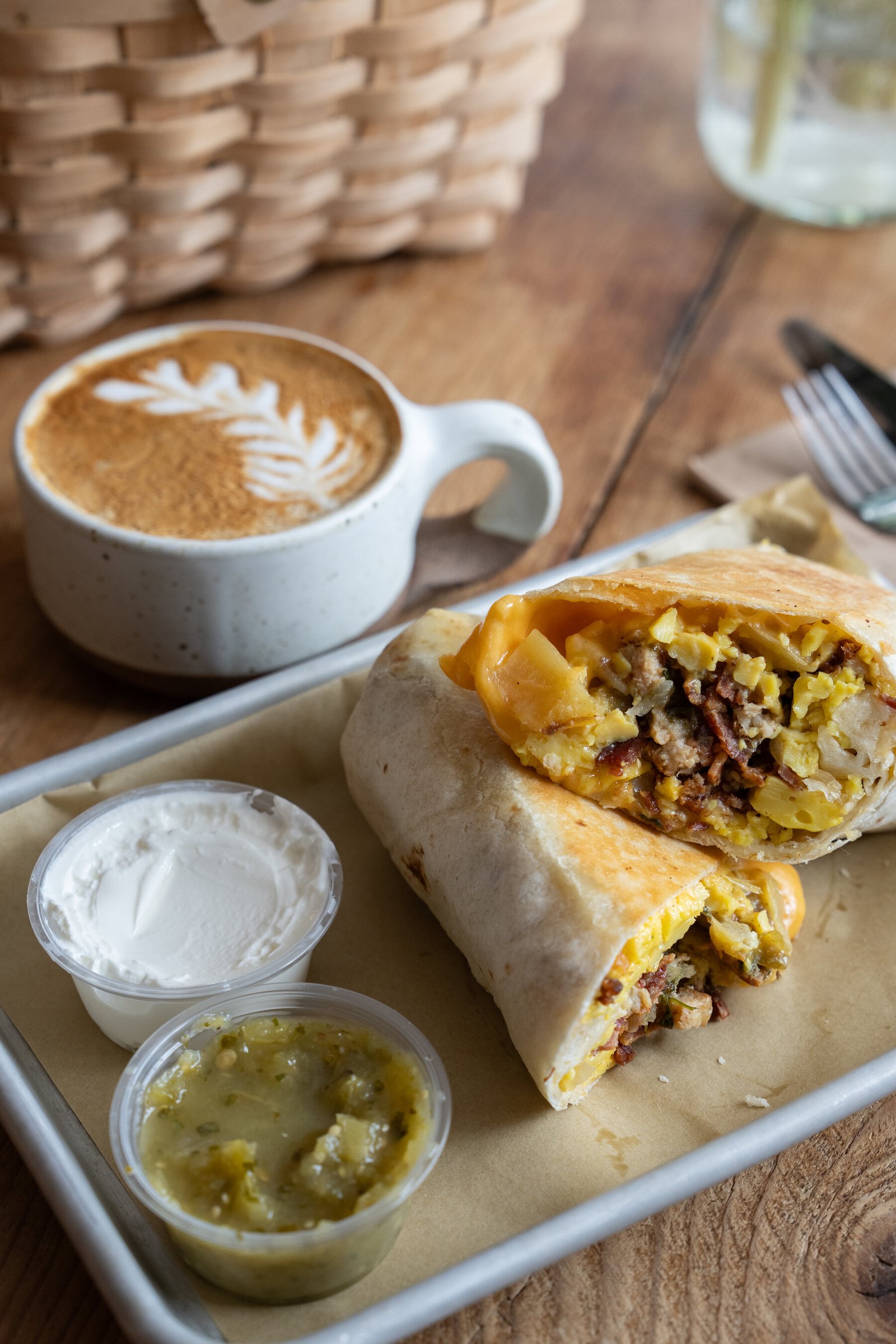 A breakfast close-up: a potato and cheddar burrito sits on brown paper on a steel tray, condiments next to it; a coffee drink is also on the table. 