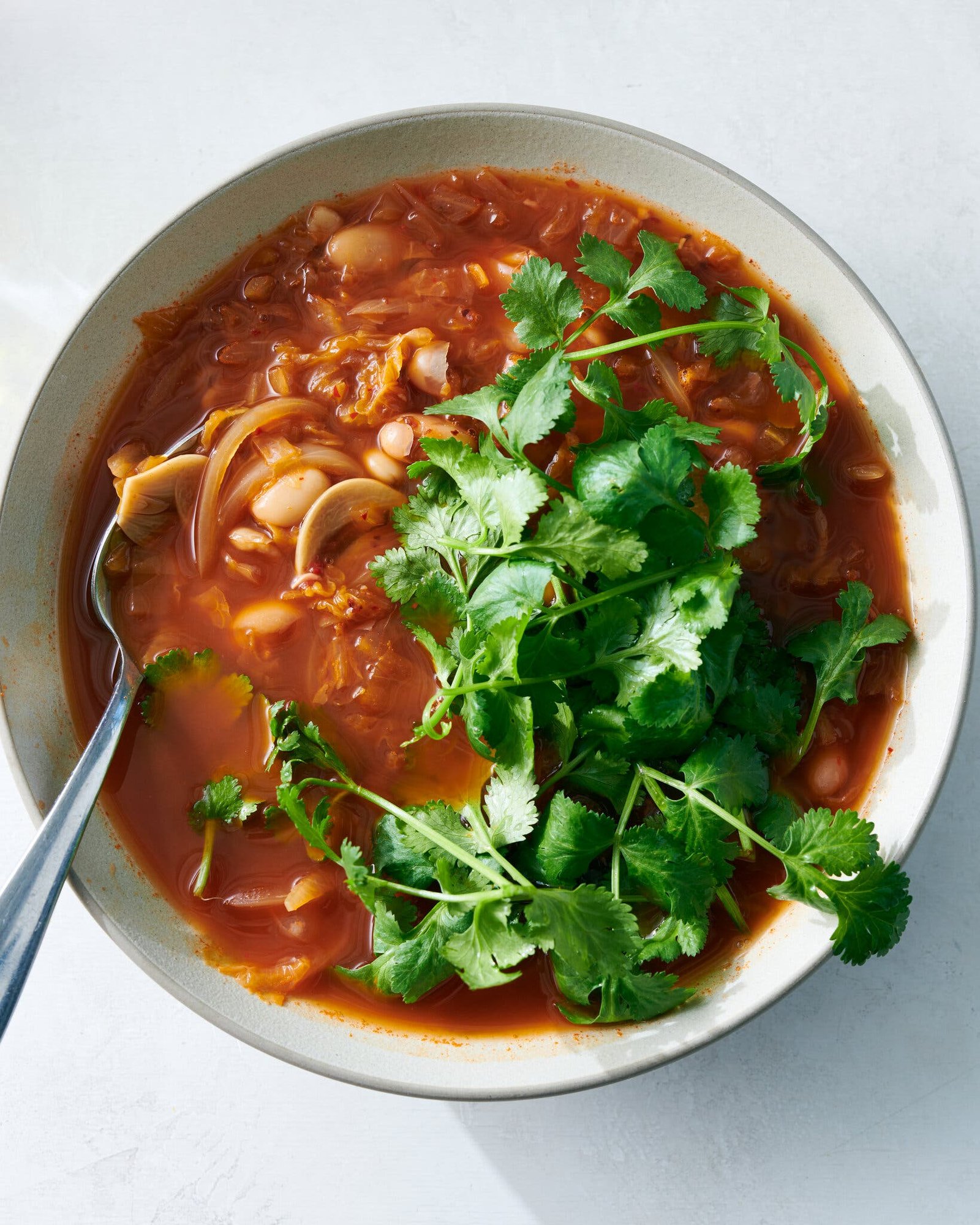 A bowl of bright red soup with beans and a leafy blanket of green cilantro, with a silver spoon.