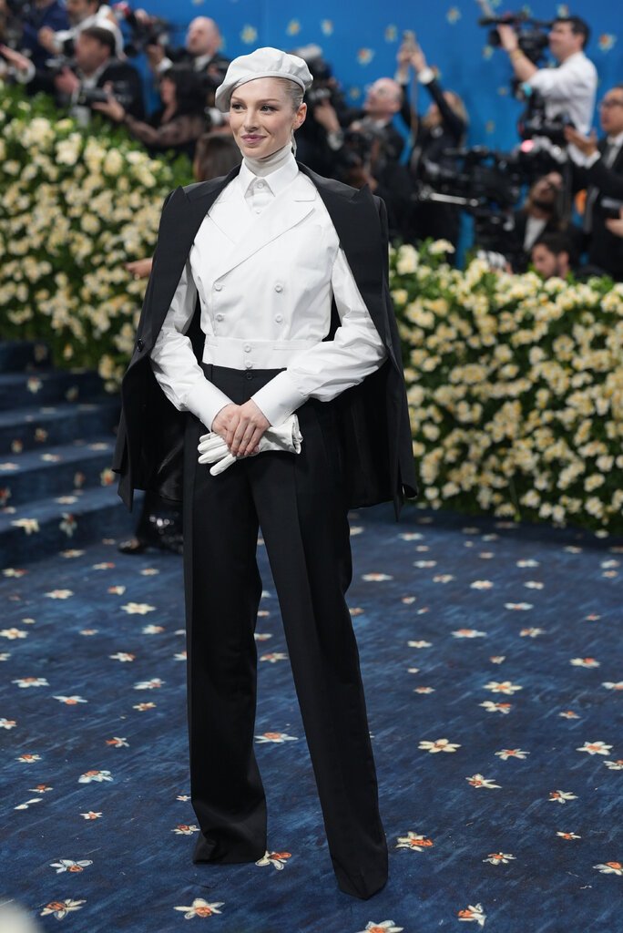 Hunter Schafer wearing black pants, a white buttoned top and white page boy cap.
