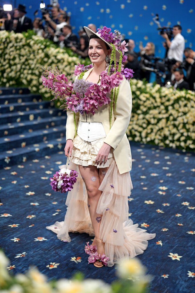 Jessica Kayll is wearing a creme suit jacket, corset, hat and ruffled skirt adorned with purple flowers.