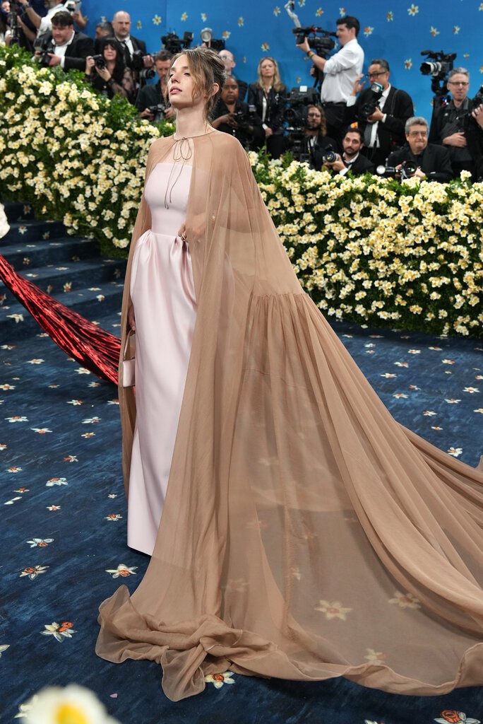 Maya Hawke wearing a blush pink gown with a sheer, floor-length beige cape tied at the neck.