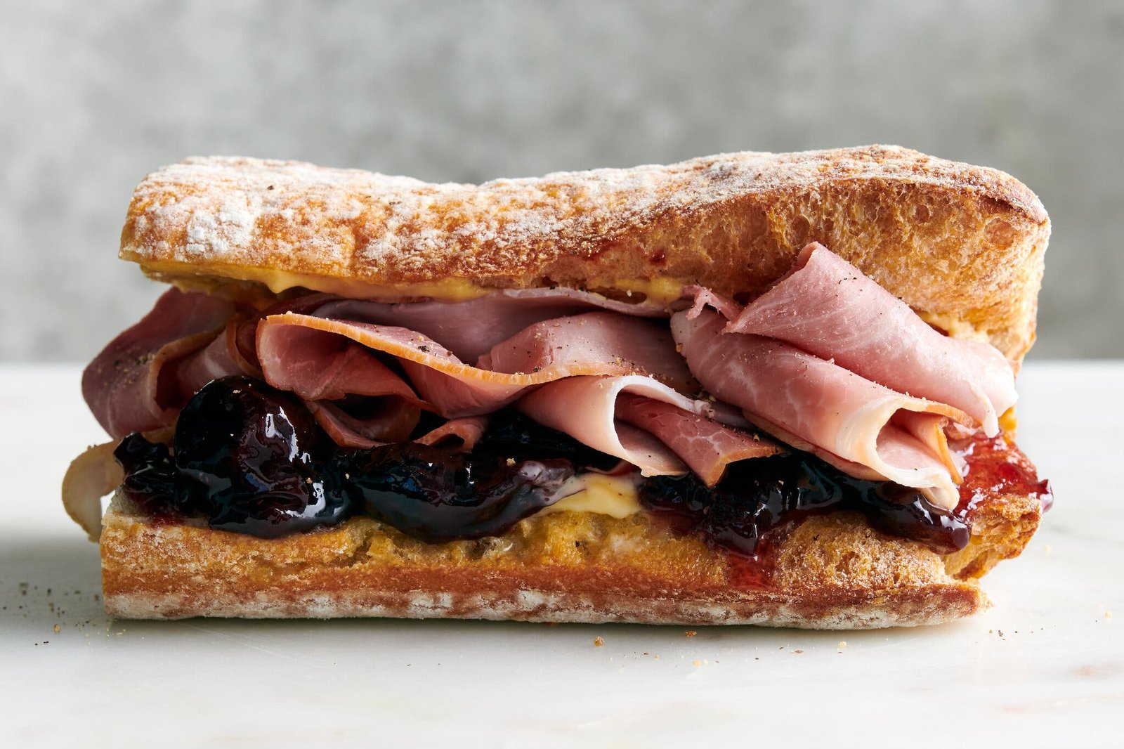 A side image of a baguette cut in half and filled with ham, rich cherry jam and butter.
