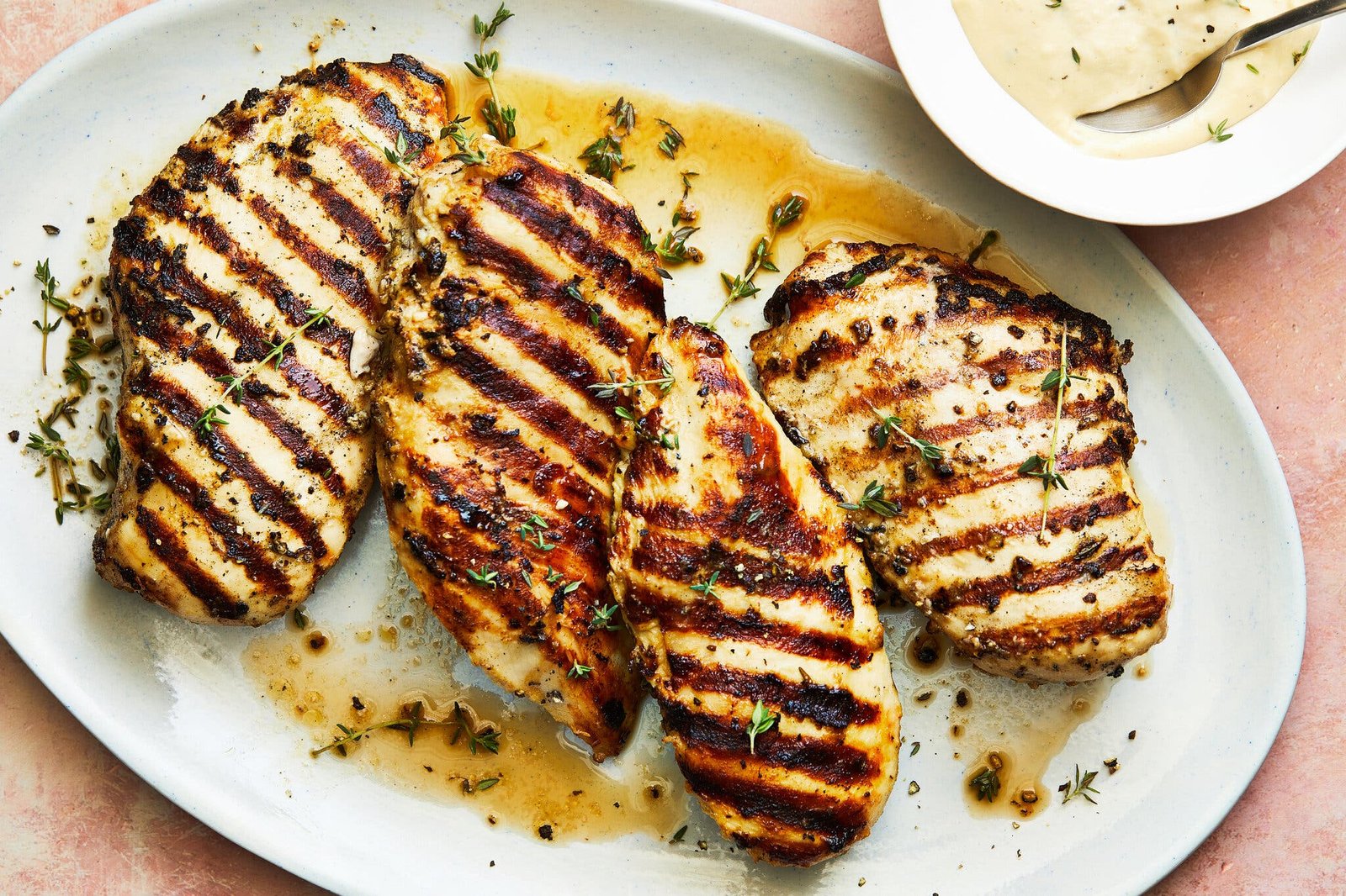 An overhead image of chicken breast finished with grill marks on a white platter. Thyme stems are scattered on top, and a sauce encircles the breasts.