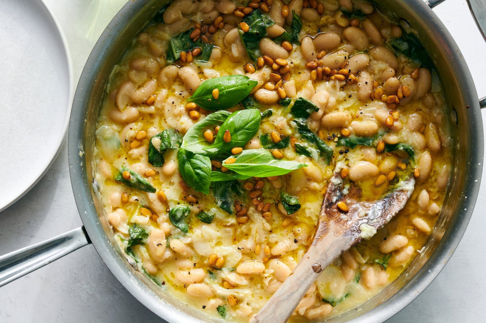A skillet holds stewed cannellini beans garnished with fresh basil and pine nuts. A wooden spoon is scooping some of the beans.