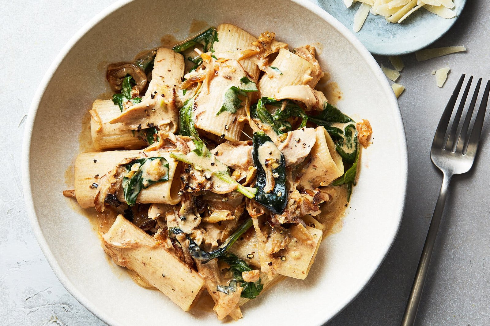 A creamy bowl of pasta topped with greens sits in a bowl. Shaved Parmesan is visible on the side.